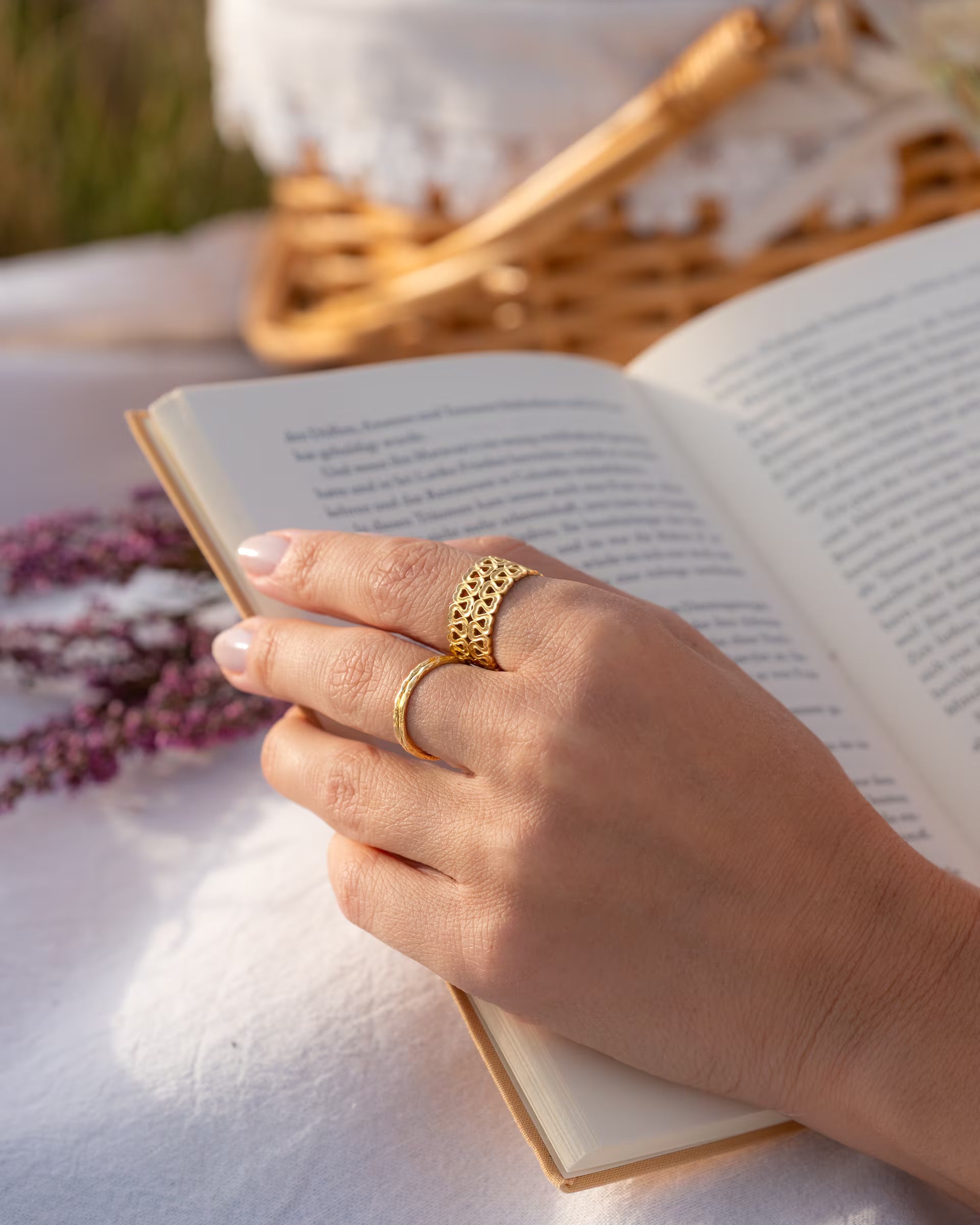 Nahaufnahme im romantischen Stil von einer Hand, die die Seiten eines geöffneten Buches hält und eine Kombination aus dem auf Bali handgefertigten goldenen, offenen ENDLESS LOVE RING mit ausgestanztem Herz-Muster sowie dem feinen SARI STACKING RING aus 18k vergoldetem 925er Silber von der Schmuckmarke ILUH trägt, im Hintergrund ein Picknickkorb und lila Heidekraut.