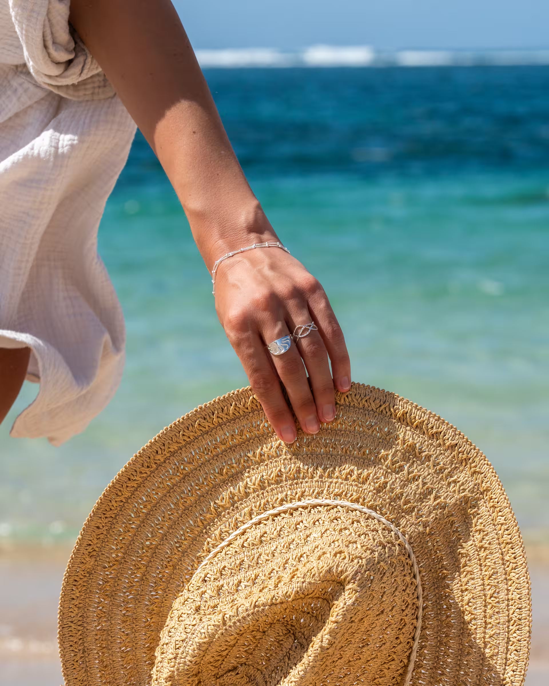 Handgefertigter silberner ARUNA Sunrise Ring, Lotus Ring und das passende Chakra Armband aus 925er Silber von der Schmuckmarke ILUH, getragen an Hand und Handgelenk einer Frau, die einen beigen Strohhut vor dem blauen Meer hält.
