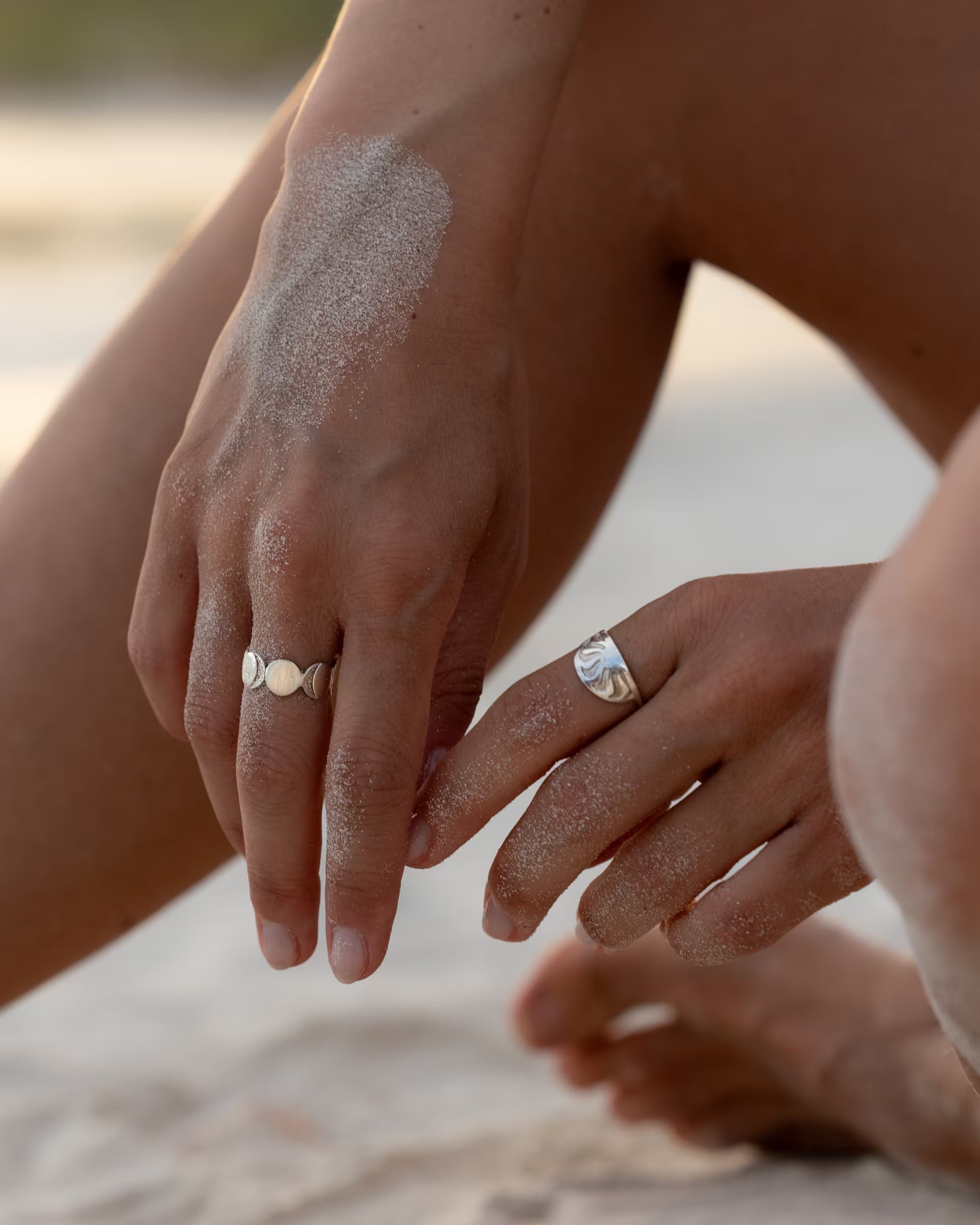 Nahaufnahme im sommerlichen Beach-Look von zwei sandigen Händen im Strandsand, die den auf Bali handgefertigten ARUNA SUNRISE Ring mit Sonnen-Motiv und den CHANDRA MOONPHASES Ring mit Mondphasen-Design aus 925er Silber von der Schmuckmarke ILUH tragen.