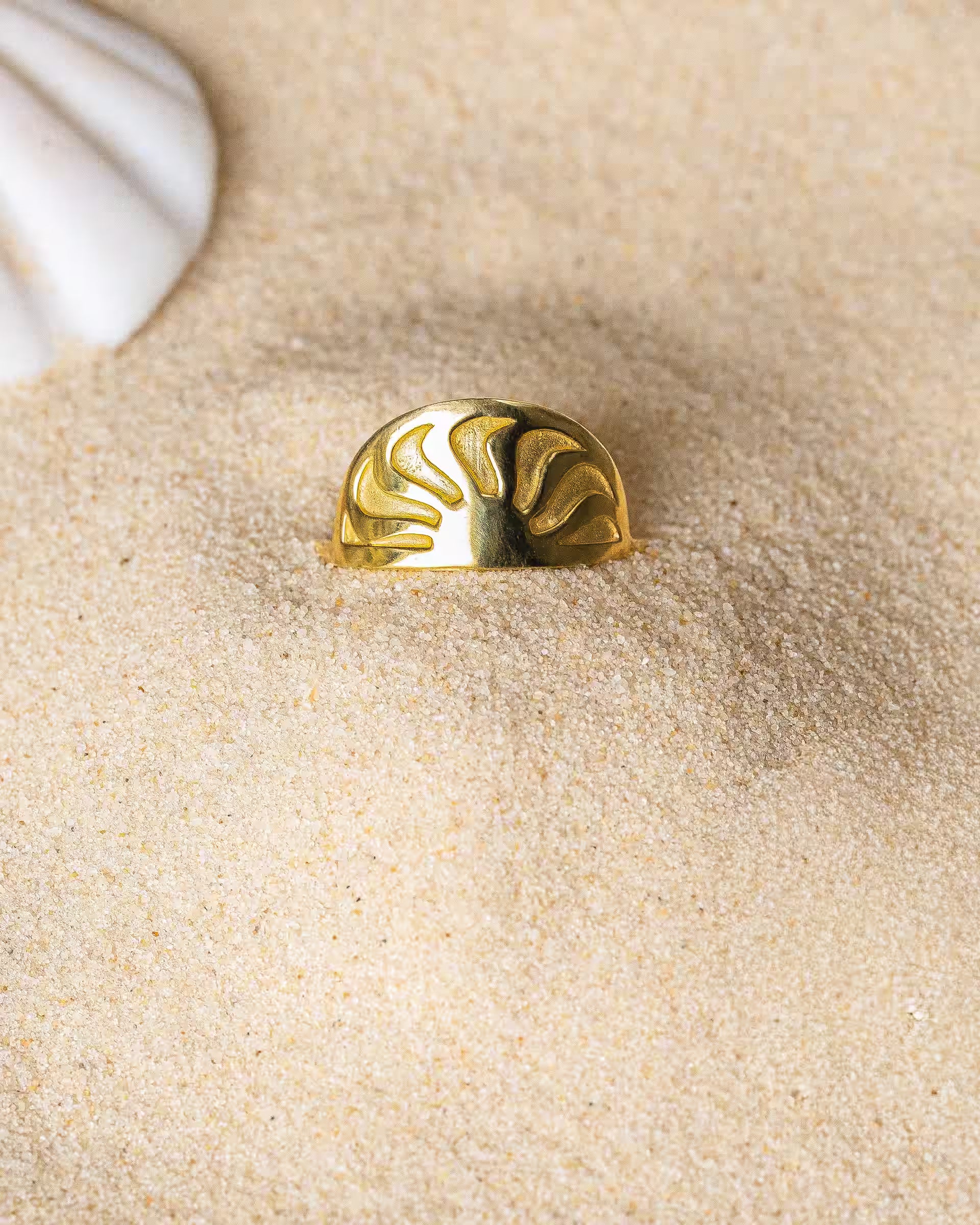 SUNRISE RING GOLD, goldener Schmuck von ILUH, handgefertigt auf Bali – Elegantes Produktfoto im maritimen Beach-Look von dem halbrunden SUNRISE RING aus 18k vergoldetem 925er Silber mit einem fein eingeprägten Sonnenstrahlen-Motiv, der dekorativ im feinen Sandstrand neben einer weißen Muschel präsentiert wird.