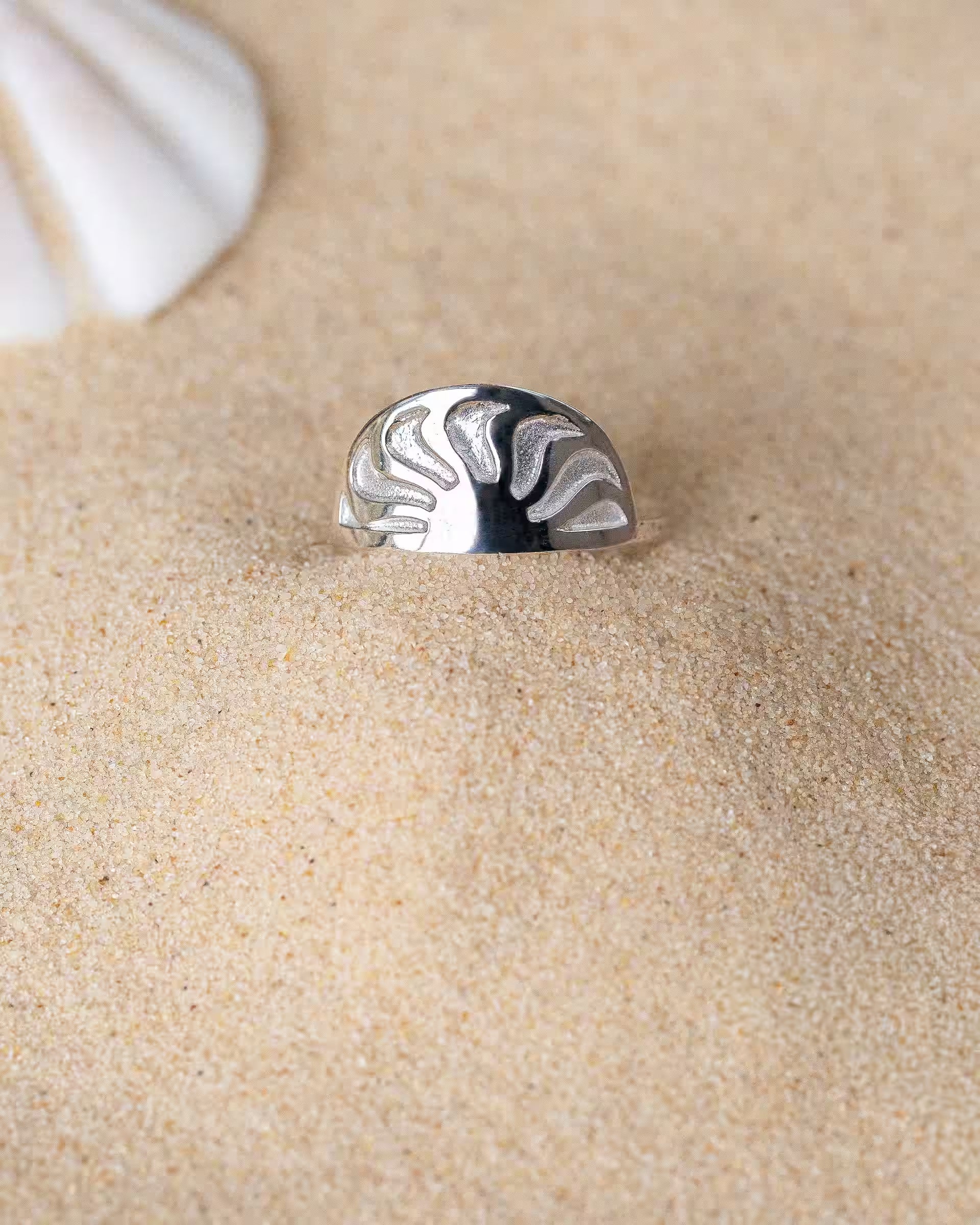 SUNRISE RING SILBER, silberner Schmuck von ILUH, handgefertigt auf Bali – Elegantes Produktfoto im maritimen Beach-Look von dem halbrunden SUNRISE RING aus 925er Silber mit einem fein eingeprägten Sonnenstrahlen-Motiv, der dekorativ im feinen Sandstrand neben einer weißen Muschel präsentiert wird.