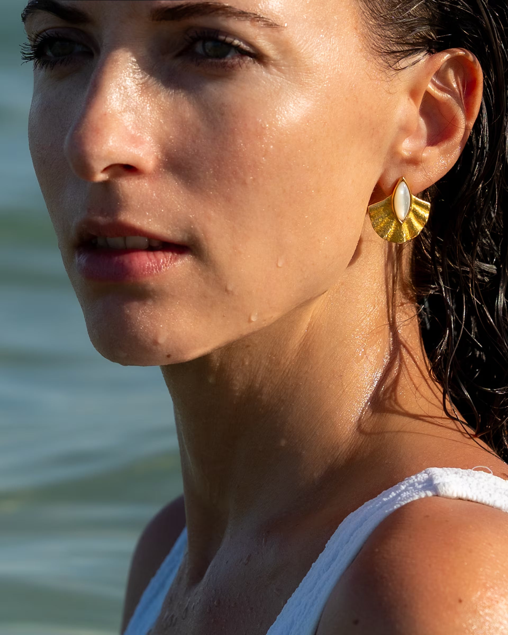 Auf Bali handgefertigte goldene ALUN Perlmutt-Ohrringe im fächerförmigen Muschel-Design aus 18k vergoldetem 925er Silber von der Schmuckmarke ILUH, getragen von einer jungen Frau auf Bali im weißen Badeanzug mit nassen Haaren, die vor dem Wasser in die Ferne blickt.