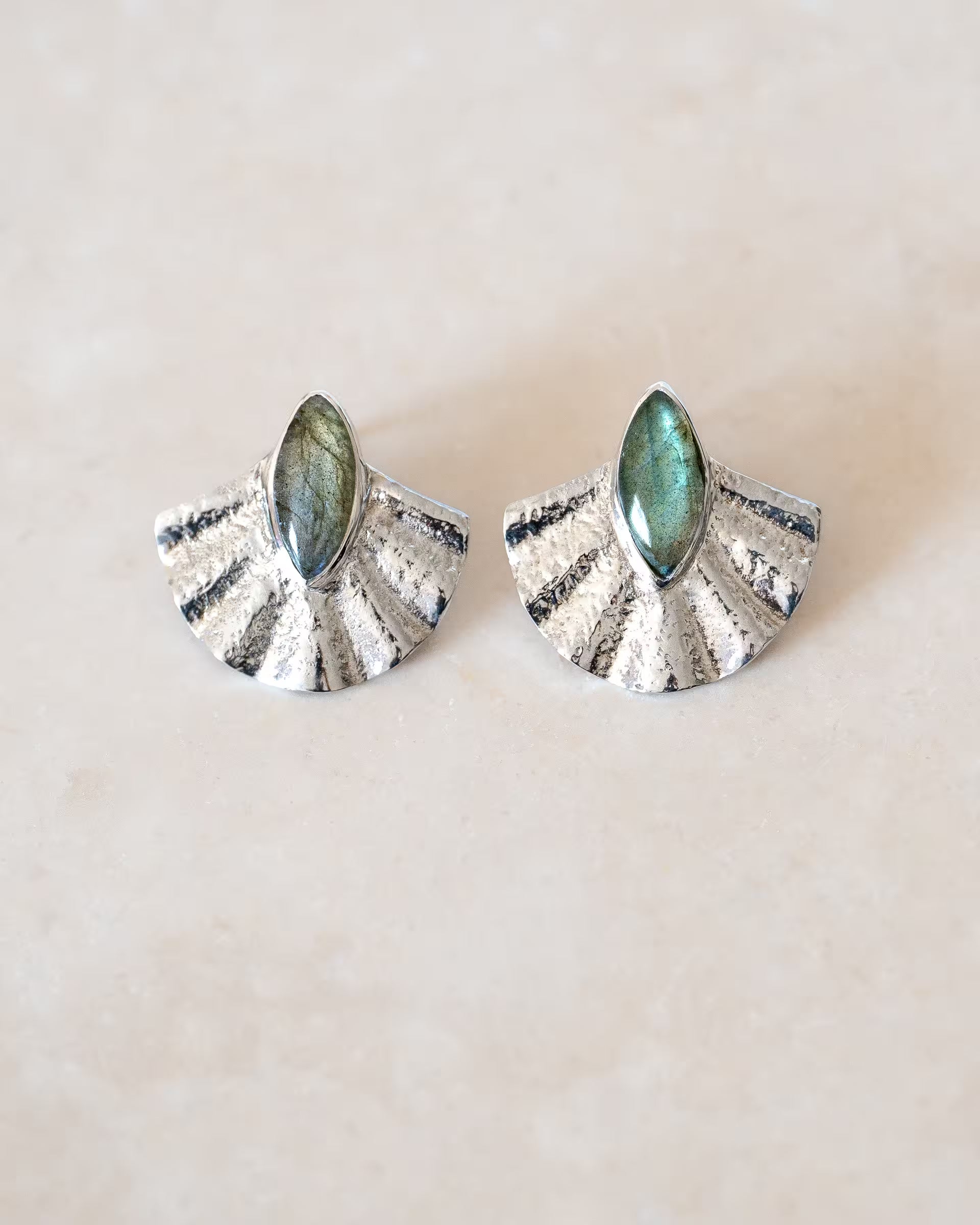 ALUN SHELL LABRADORITE OHRRINGE SILBER, silberne Ohrstecker von ILUH, handgefertigt auf Bali – Stilvolles Produktfoto im minimalistischen Boho-Look von einem Paar fächerförmiger Ohrstecker aus 925er Silber mit einer reliefartigen Struktur und je einem mittig gefassten, marquise-förmigen Labradorit-Edelstein, der intensiv grünblau schimmert, flach präsentiert auf einem hellen Naturstein-Untergrund.