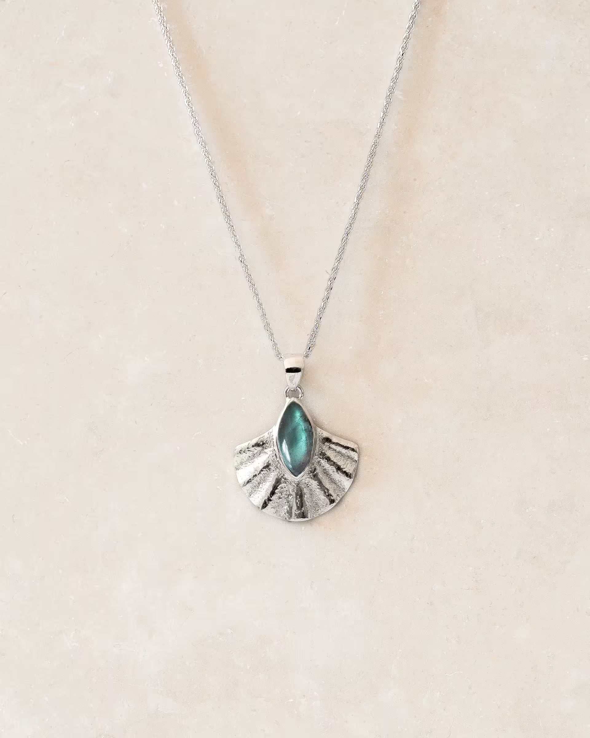 ALUN SHELL LABRADORITE KETTE SILBER, silberne Halskette von ILUH, handgefertigt auf Bali – Elegantes Produktfoto im minimalistischen Boho-Look von einer feinen Halskette aus 925er Silber mit einem fächerförmigen Anhänger, der einen intensiv blaugrün schimmernden Labradorit-Edelstein im Cabochonschliff zeigt, flach präsentiert auf einem hellen, neutralen Steinuntergrund.