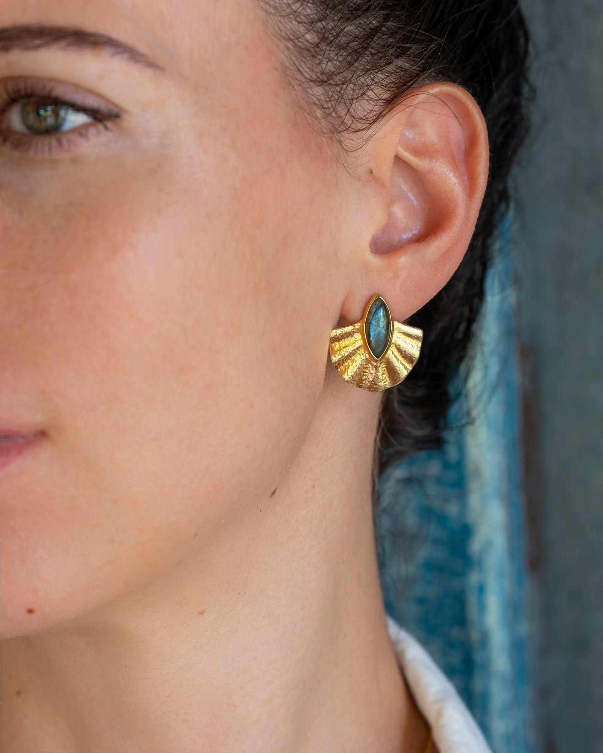 ALUN SHELL LABRADORITE OHRRINGE, goldener Schmuck von ILUH, handgefertigt auf Bali – Stilvolle Nahaufnahme im Profil von einer Frau, die fächerförmige Ohrringe aus 18k vergoldetem 925er Silber mit einem intensiv blaugrün schimmernden Labradorit-Edelstein im Cabochonschliff vor einem rustikalen, blauen Holzhintergrund präsentiert.