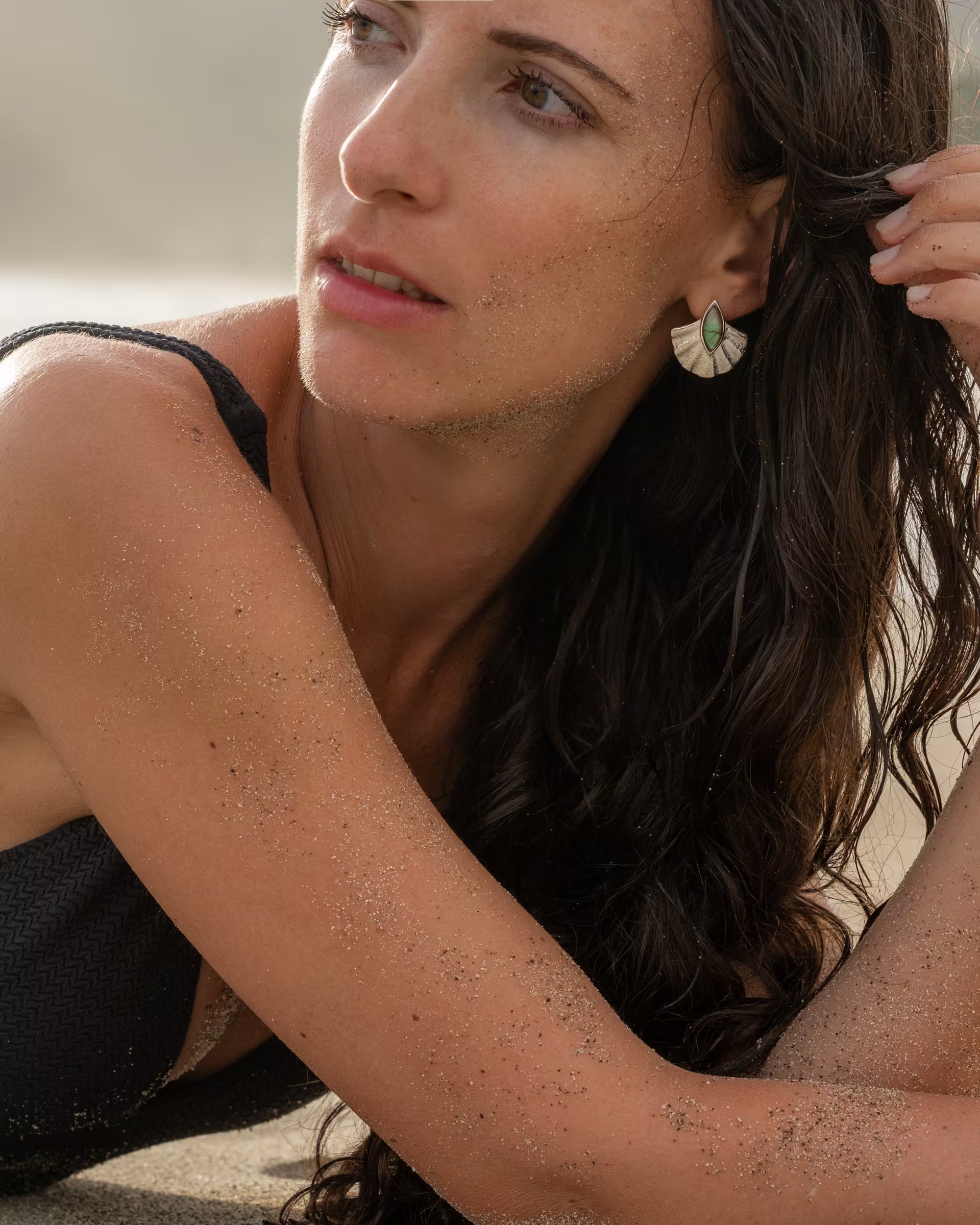 Im warmen, natürlichen Licht des Küstenhimmels liegt das Modell mit sanft von feinen Sandkörnern verzierten Armen elegant am Strand, während ihr verträumter Blick die auf Bali handgefertigten silbernen ALUN SHELL OHRRINGE aus 925er Sterlingsilber perfekt in Szene setzt, deren detailreiches Fächer-Design wunderschön mit dem tiefgrünen Labradorit-Edelstein kontrastiert.