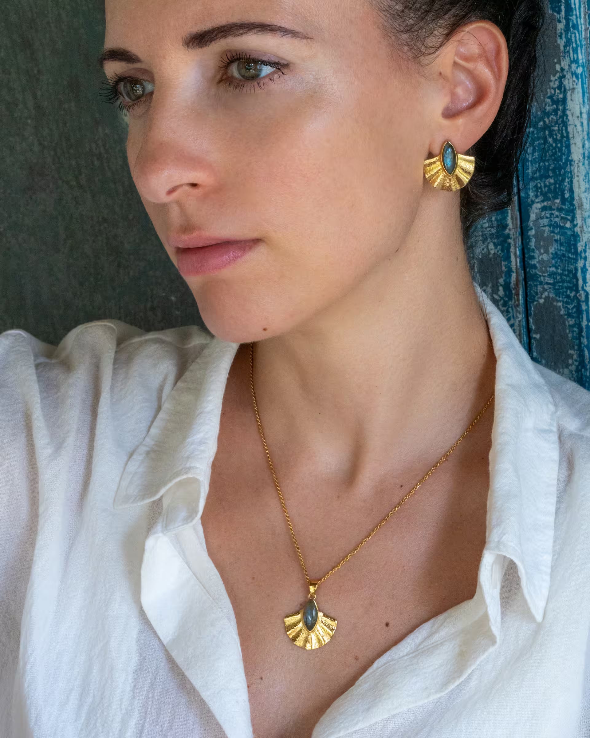ALUN SHELL GOLD SET, goldene Halskette und Ohrringe von ILUH, handgefertigt auf Bali – Stilvolle Nahaufnahme im Profil von einer wegschauenden Frau im weißen Leinenhemd vor einer rustikalen, blauen Holzwand, die eine filigrane Kette und die passenden fächerförmigen Ohrringe aus 18k vergoldetem 925er Silber mit blau-grün schimmernden Labradorit-Edelsteinen präsentiert.