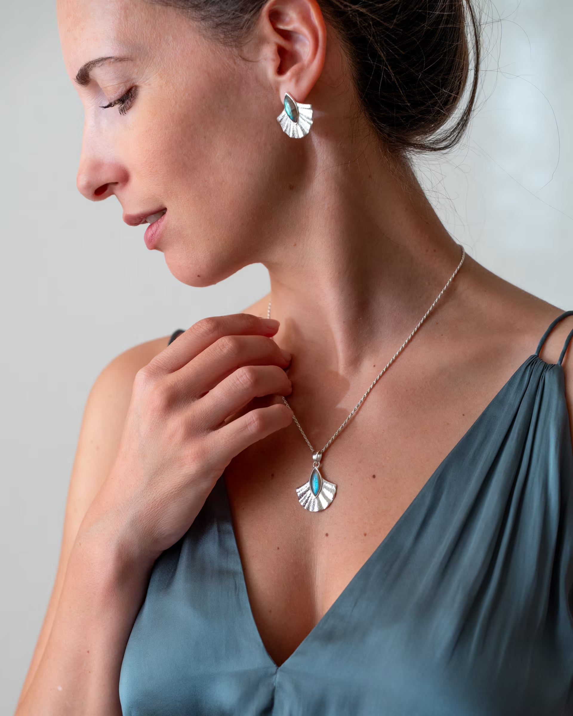 ALUN SHELL SILBER SET, silberne Halskette und Ohrringe von ILUH, handgefertigt auf Bali – Elegante Nahaufnahme im Profil von einer lächelnden Frau im seidigen, blaugrauen Spaghettiträgertop vor hellem Hintergrund, die eine Hand zart an ihre feine Halskette legt und das passende fächerförmige Schmuckset aus 925er Silber mit türkis-blau schimmernden Labradorit-Edelsteinen zeigt.