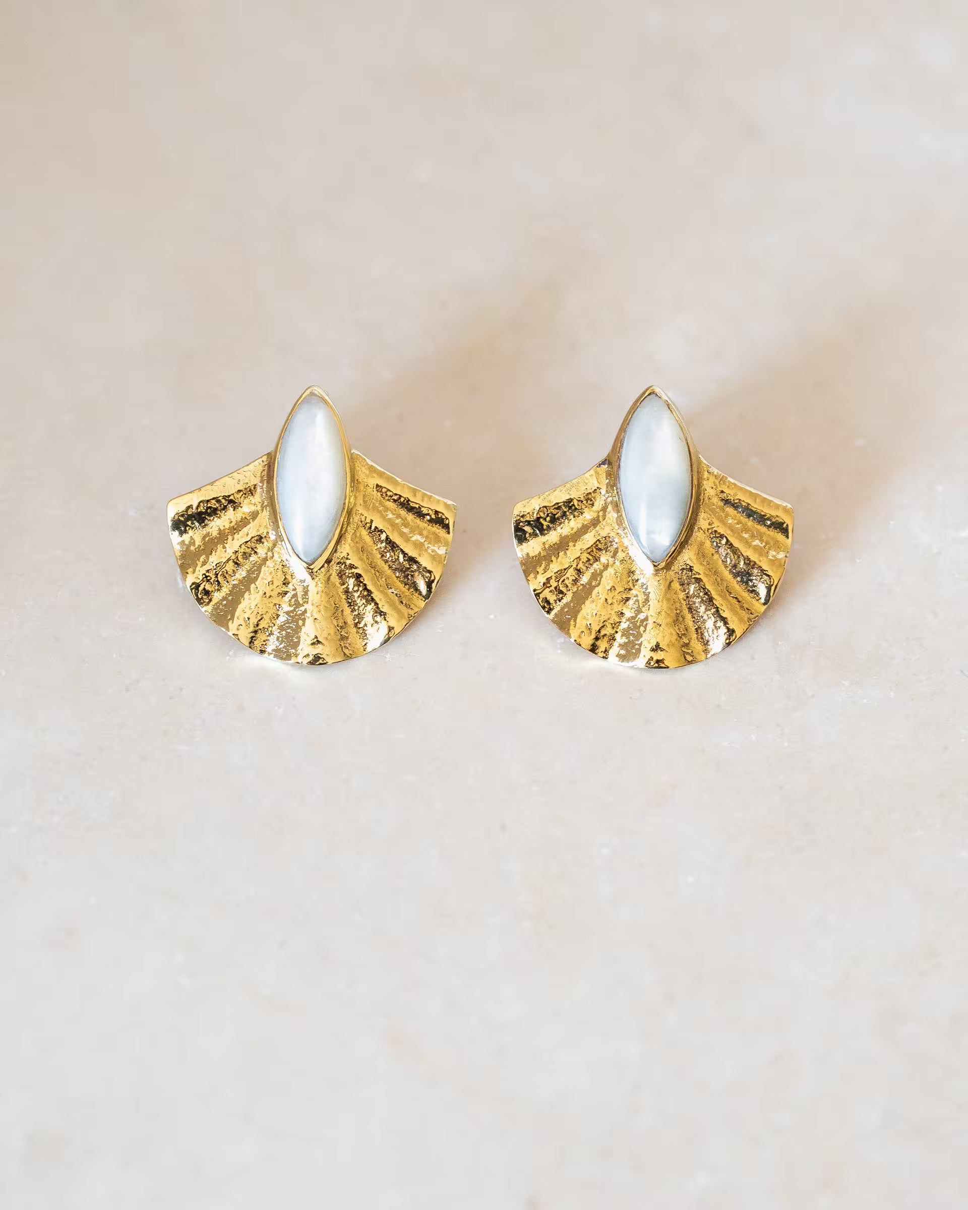 ALUN SHELL MOTHER OF PEARL OHRRINGE GOLD, goldene Ohrstecker von ILUH, handgefertigt auf Bali – Stilvolles Produktfoto im minimalistischen Boho-Look von einem Paar fächerförmiger Ohrstecker aus 18k vergoldetem 925er Silber mit einer reliefartigen Struktur und je einem mittig gefassten, marquise-förmigen Perlmutt-Stein, flach präsentiert auf einem hellen Naturstein-Untergrund.