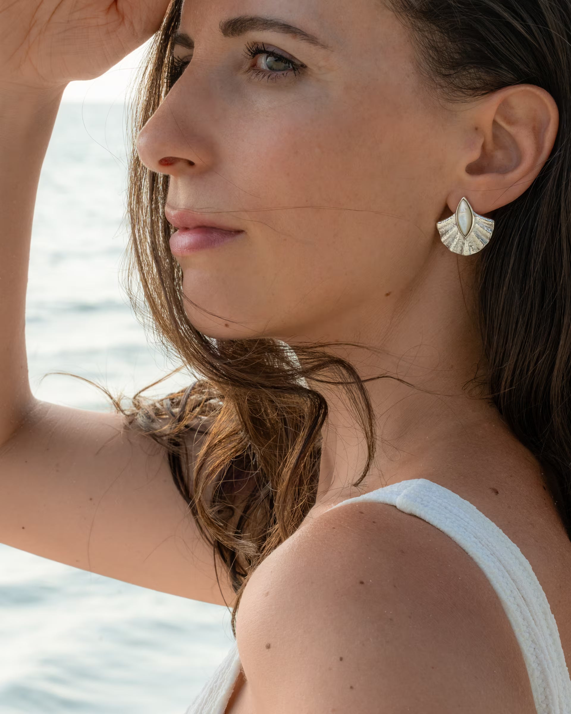 Den Blick entspannt in die Ferne über das glitzernde Wasser gerichtet, schützt das Modell im weißen Oberteil die Augen vor der Sonne und unterstreicht ihren maritimen Beach-Look mit den auf Bali handgefertigten silbernen ALUN SHELL OHRRINGEN aus 925er Silber, die mit edlem, weißem Perlmutt verziert sind.