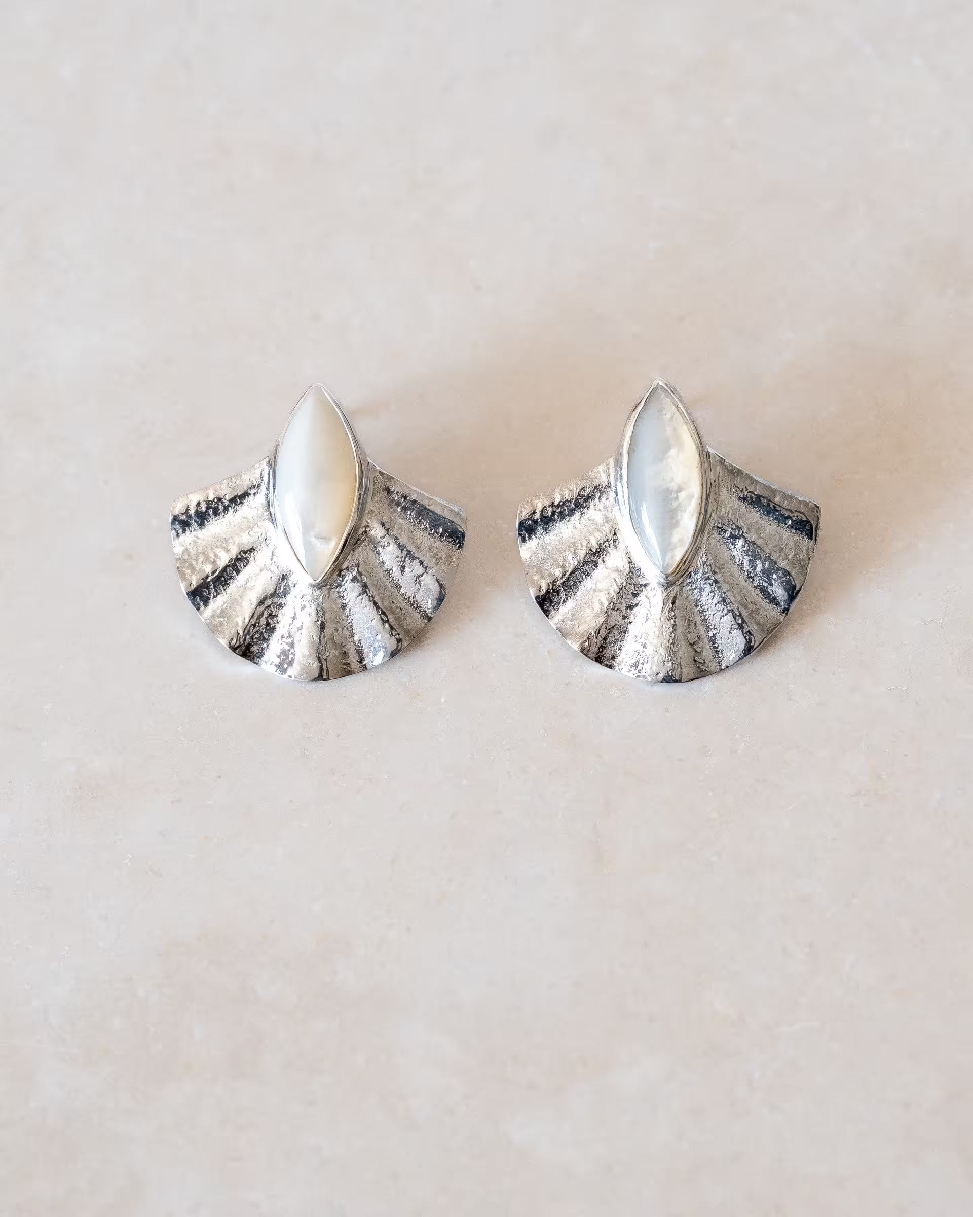 ALUN SHELL MOTHER OF PEARL OHRRINGE SILBER, silberne Ohrstecker von ILUH, handgefertigt auf Bali – Stilvolles Produktfoto im minimalistischen Boho-Look von einem Paar fächerförmiger Ohrstecker aus 925er Silber mit einer reliefartigen Struktur und je einem mittig gefassten, marquise-förmigen Perlmutt-Stein, flach präsentiert auf einem hellen Naturstein-Untergrund.