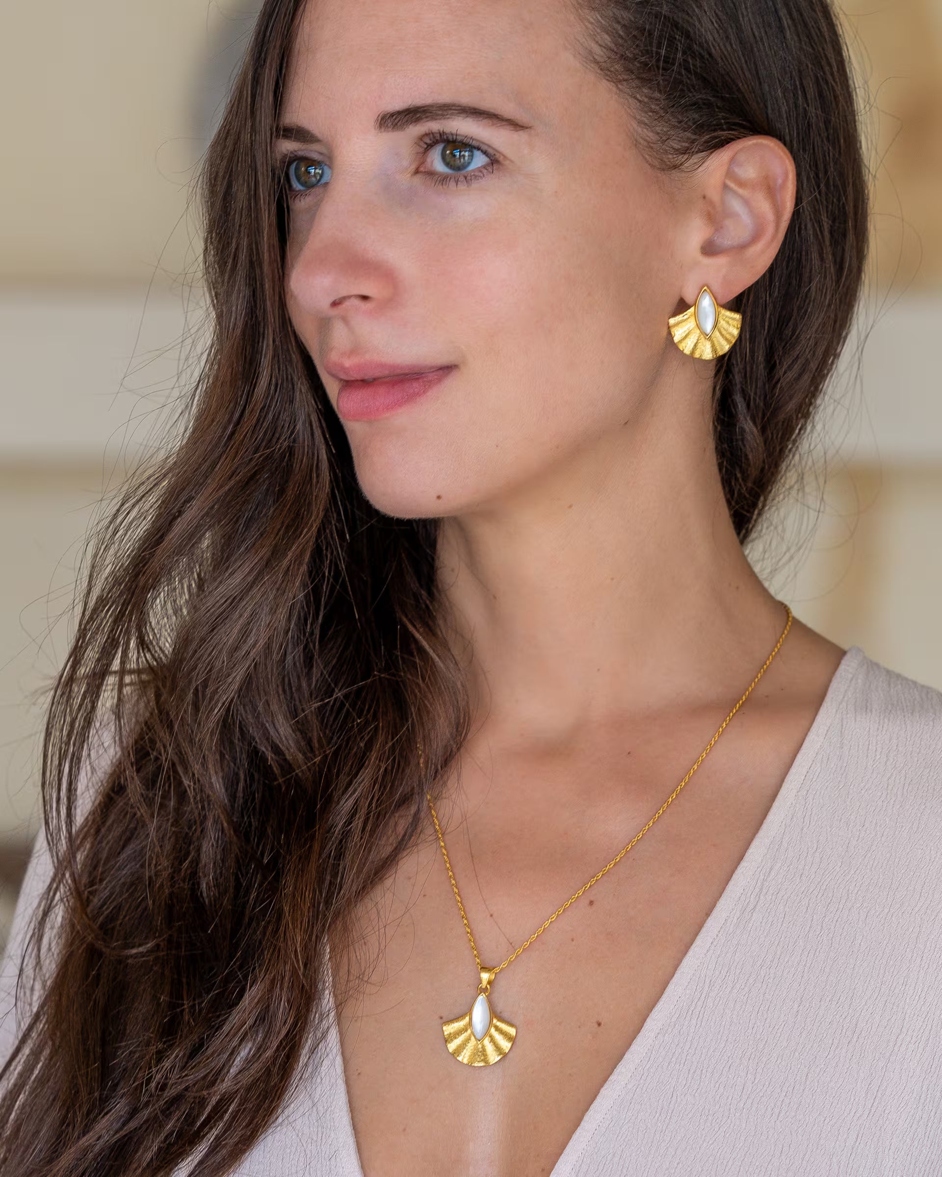Nahaufnahme im eleganten Boho-Look von einer Frau im Profil mit langen, braunen Haaren und hellem V-Ausschnitt-Oberteil, die eine Kombination aus auf Bali handgefertigtem goldenem Schmuck – bestehend aus der ALUN SHELL KETTE und den passenden ALUN SHELL OHRRINGEN mit fächerförmigen Anhängern, verziert mit schimmerndem, weißem Perlmutt – aus 18k vergoldetem 925er Silber von der Schmuckmarke ILUH trägt.