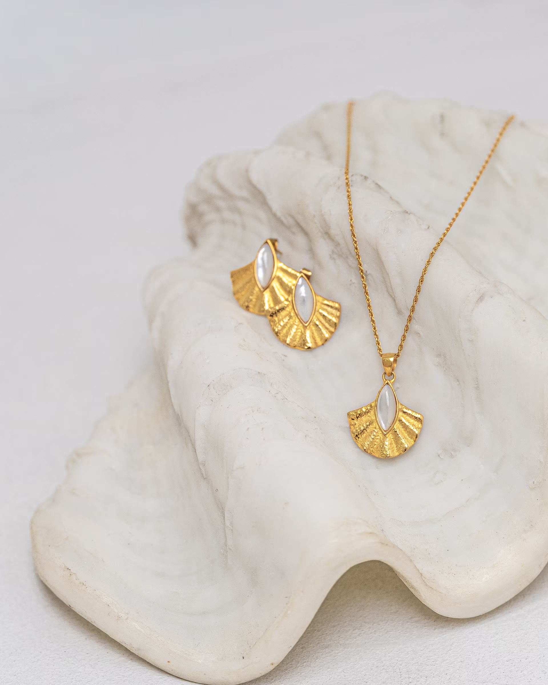 ALUN SHELL PERLMUTT SCHMUCKSET, goldene Halskette und Ohrringe von ILUH, handgefertigt auf Bali – Elegantes Produktfoto im maritimen Look von einem feinen Schmuckset aus 18k vergoldetem 925er Silber mit fächerförmigen Anhängern und weißer Perlmutt-Einlage, das dekorativ auf einer großen, strukturierten weißen Muschel vor hellem Hintergrund präsentiert wird.
