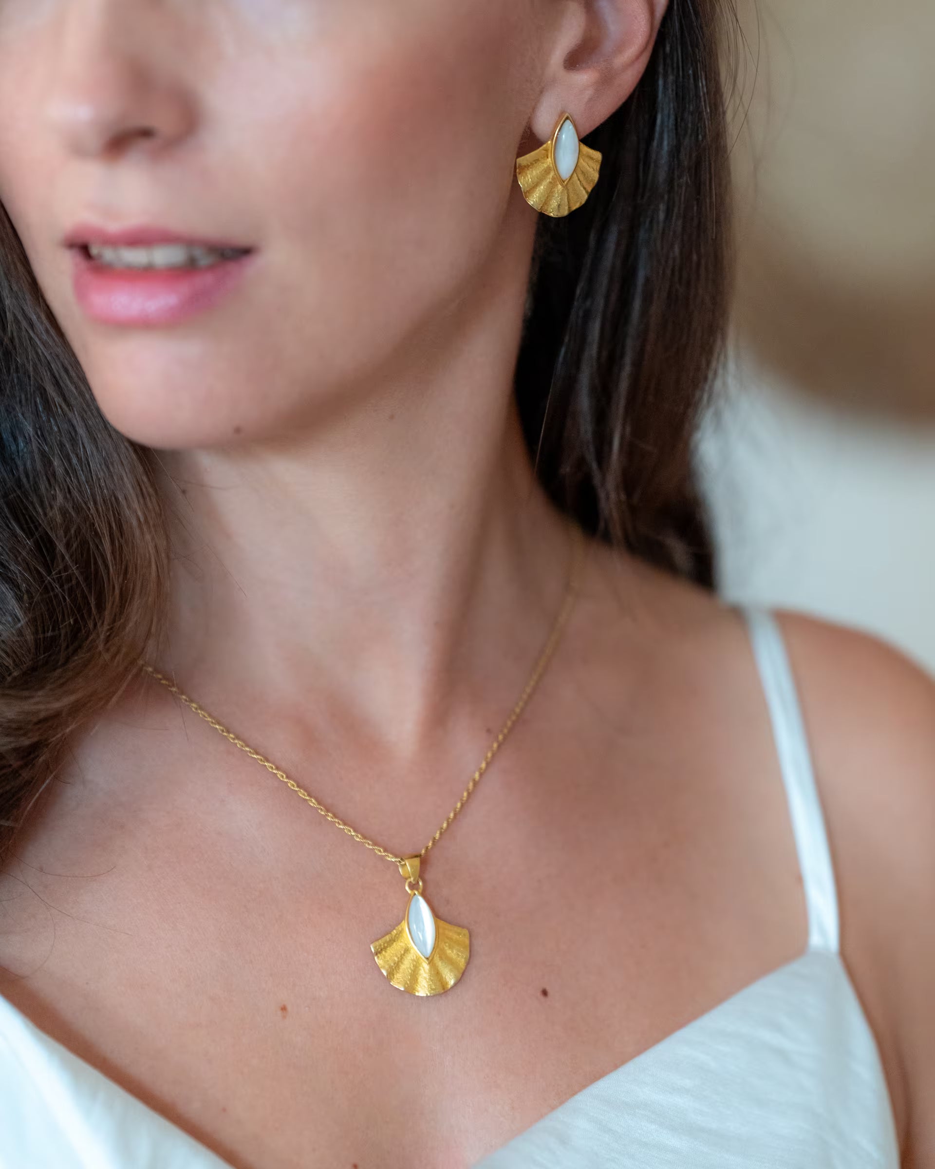 ALUN SHELL GOLD SET, goldene Halskette und Ohrringe von ILUH, handgefertigt auf Bali – Stilvolle Nahaufnahme von einer Frau im weißen Top aus Seide, die das fächerförmige Schmuckset aus 18k vergoldetem 925er Silber mit edel glänzenden, weißen Perlmutt elegant präsentiert.