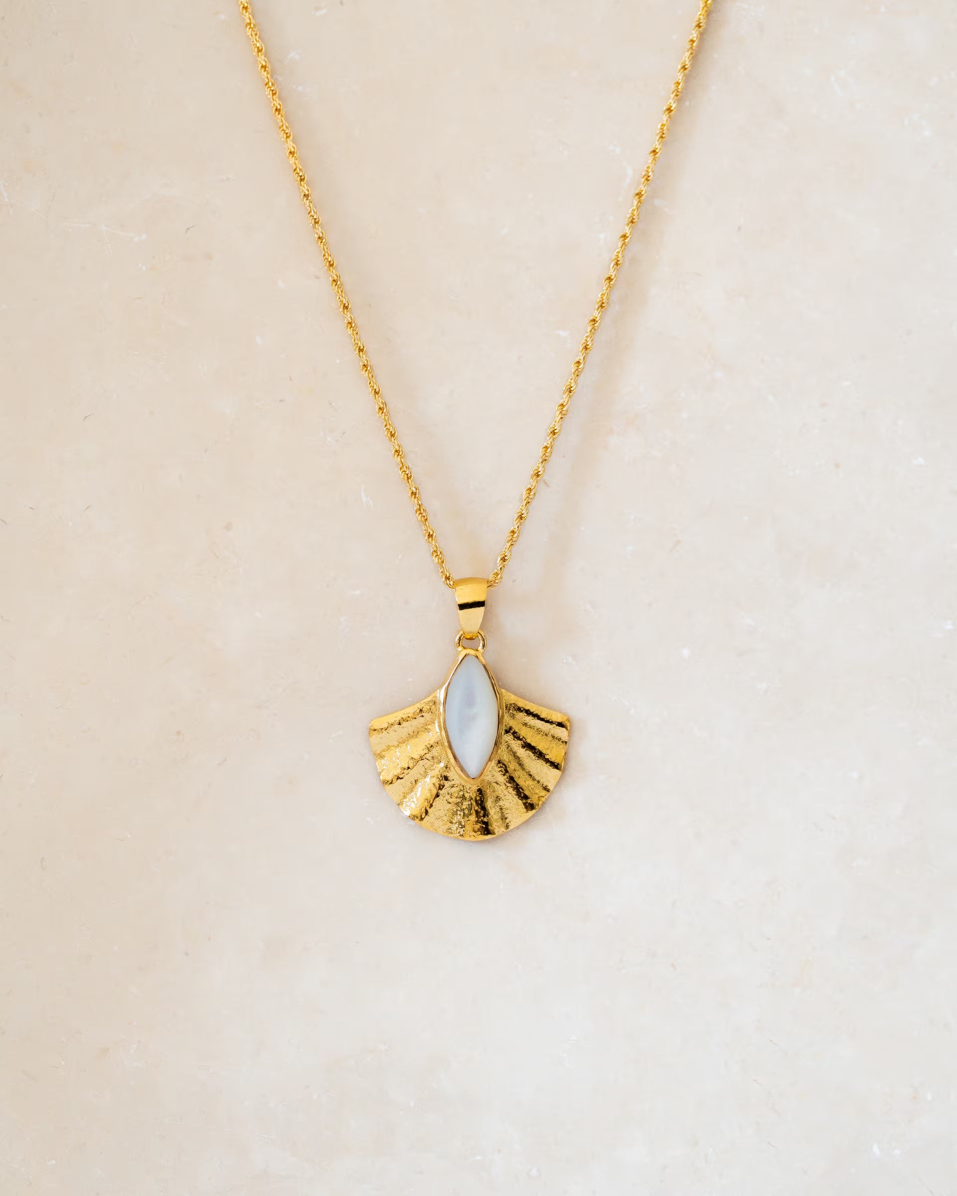 Diese handgefertigte goldene ALUN SHELL MOTHER OF PEARL HALSKETTE von der Schmuckmarke ILUH im minimalistischen Boho-Look besticht durch einen fächerförmigen Anhänger mit reliefartiger Struktur und einen mittig gefassten marquise-förmigen Perlmutt-Stein, der deinem Outfit eine magische, balinesische Note verleiht.