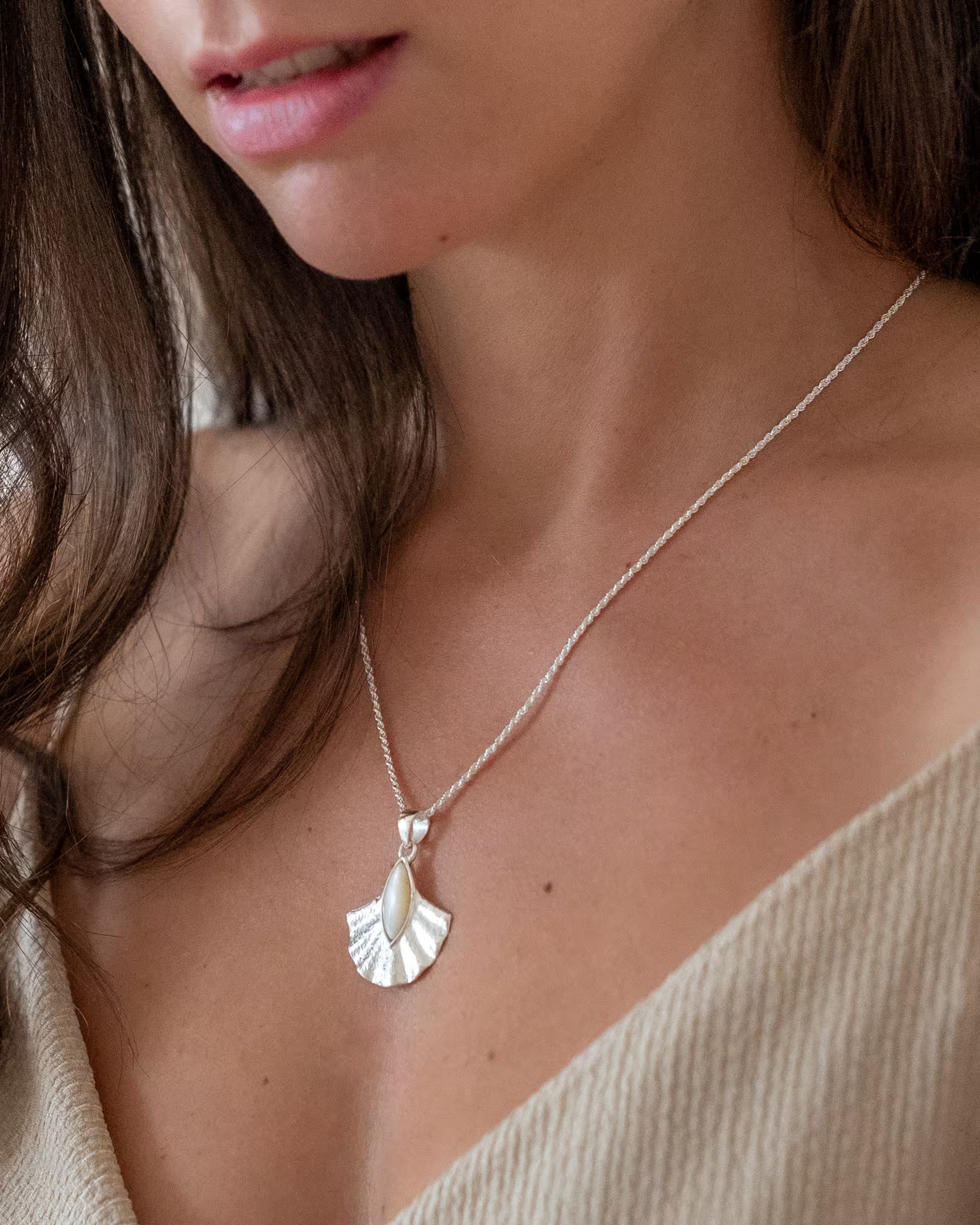 Nahaufnahme im minimalistisch-eleganten Look von dem Dekolleté einer Frau mit langen braunen Haaren im hellen Oberteil, die die auf Bali handgefertigte silberne ALUN SHELL KETTE mit einem fächerförmigen Anhänger, verziert mit schimmerndem, weißem Perlmutt, aus 925er Silber von der Schmuckmarke ILUH trägt.