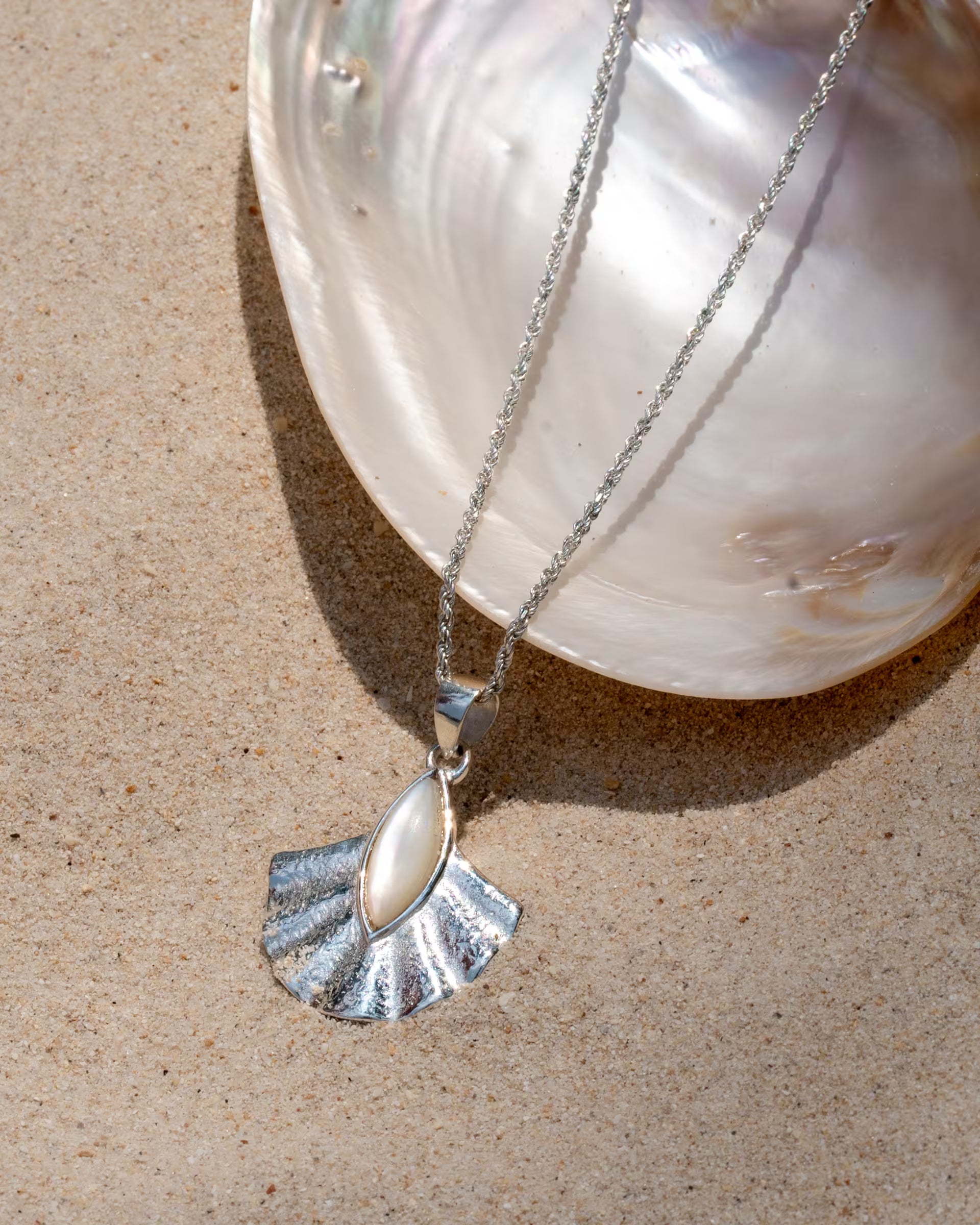 Produktfoto im maritimen Beach-Look von der auf Bali handgefertigten silbernen ALUN SHELL KETTE mit einem fächerförmigen Anhänger, der in der Mitte mit schimmerndem, weißem Perlmutt verziert ist, kunstvoll platziert auf einer großen, glänzenden Muschelschale im hellen Strandsand.