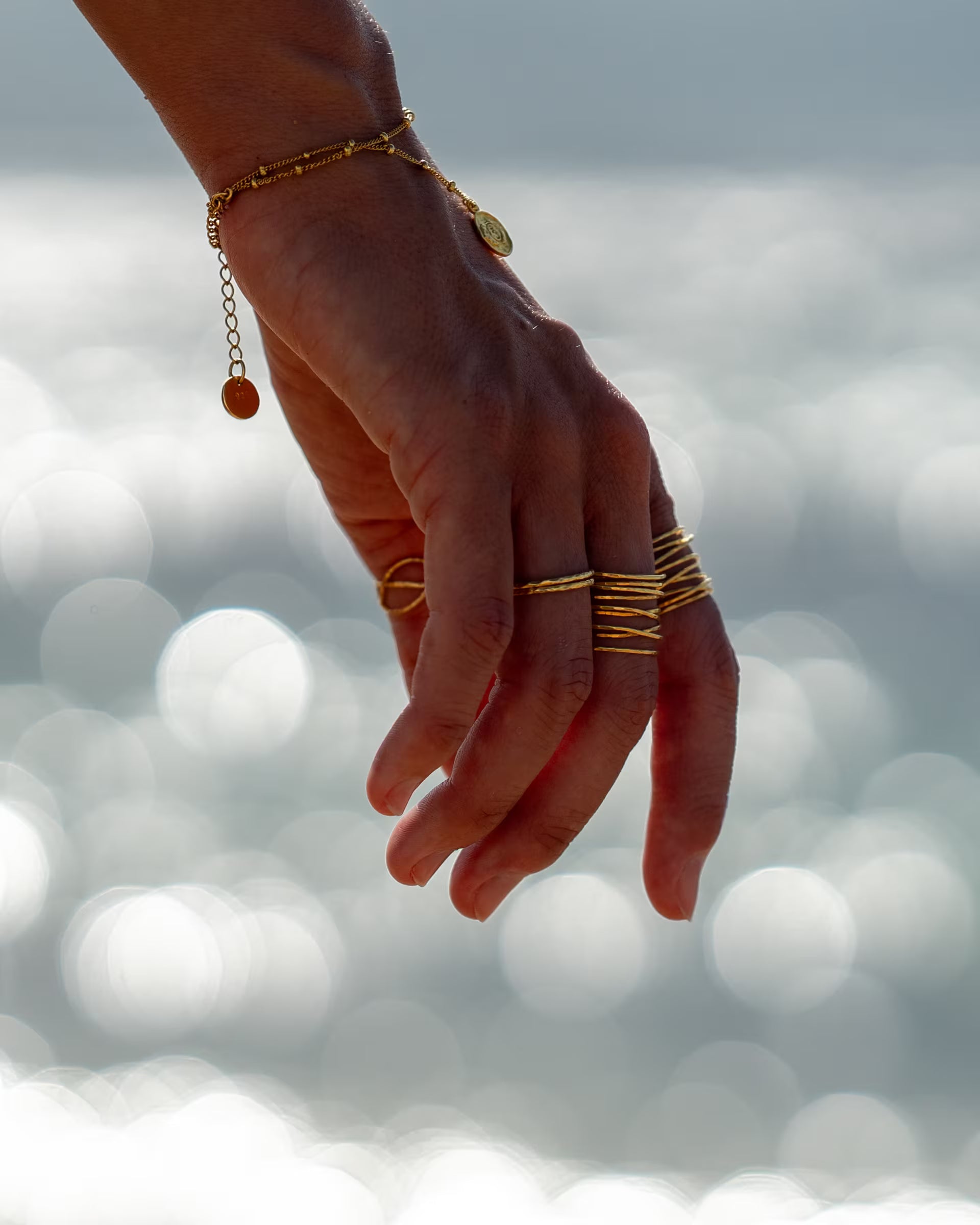 AURA RING GOLD, goldener Schmuck von ILUH, handgefertigt auf Bali – Detailreiche Nahaufnahme im maritimen Beach-Look von einer Hand, die im eleganten Gegenlicht über glitzerndem Meerwasser schwebt und dabei das filigrane Chakra-Armband sowie eine Kombination aus dem breiten, mehrreihigen, gehämmerten AURA RING im Wickeldesign und schmalen Ringen aus 18k vergoldetem 925er Silber präsentiert.