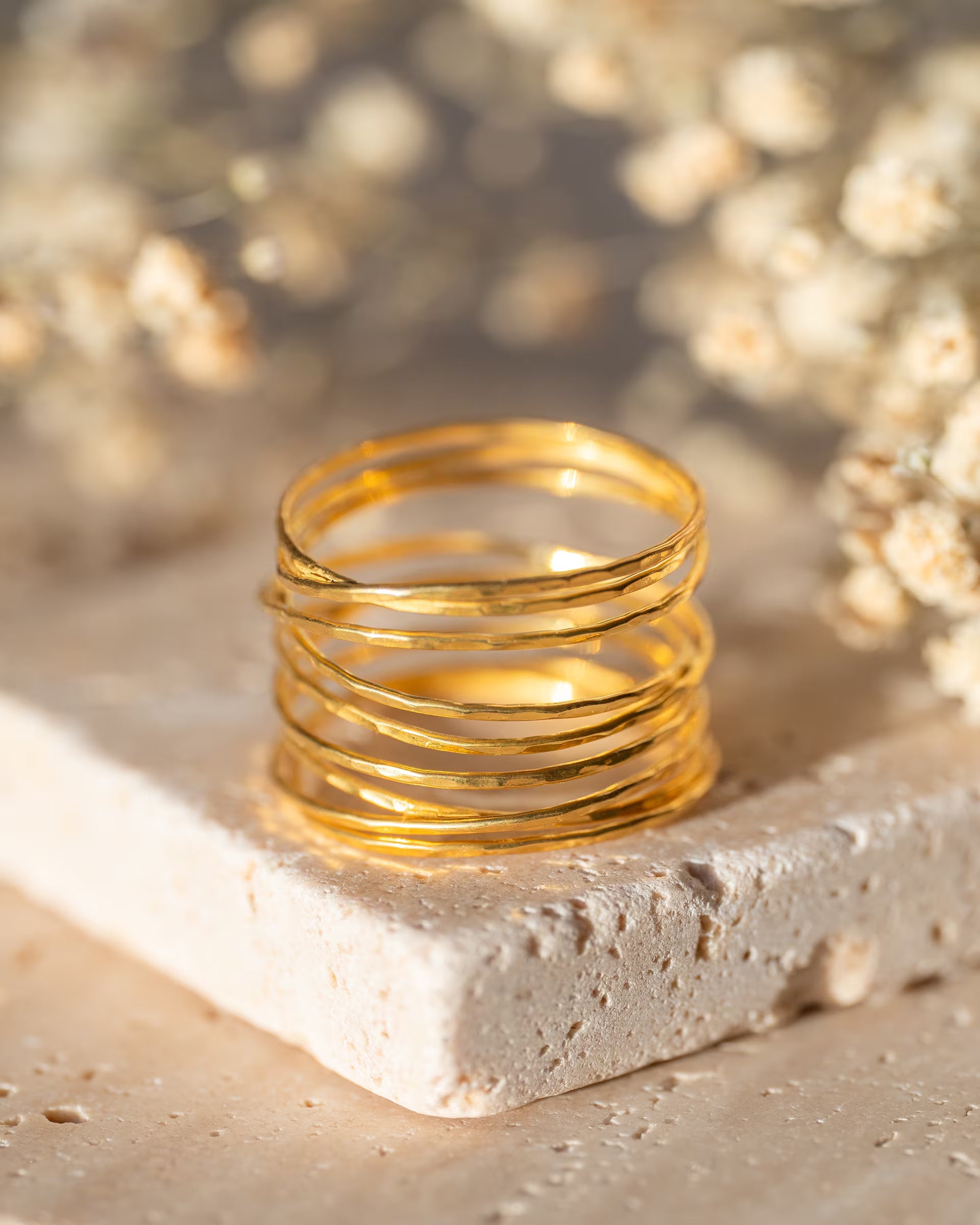 AURA RING GOLD, goldener Schmuck von ILUH, handgefertigt auf Bali – Elegantes Produktfoto im warmen Minimalist-Look von dem breiten, mehrreihigen, gehämmerten AURA RING im Wickeldesign aus 18k vergoldetem 925er Silber, der dekorativ auf einem hellen Travertin-Steinblock vor einem weichgezeichneten Hintergrund aus zarten Trockenblumen präsentiert wird.