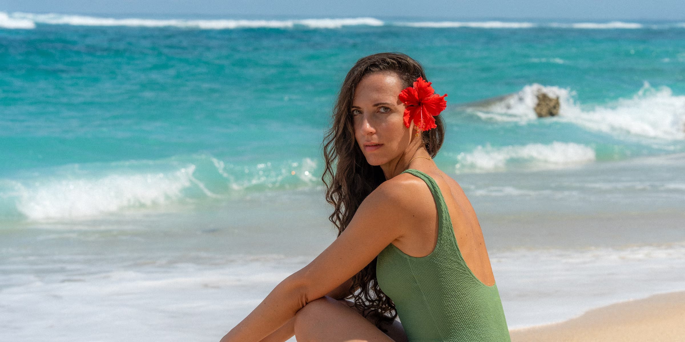 Eine junge Frau mit roter Hibiskusblüte im Haar sitzt entspannt an einem traumhaften Strand auf Bali und trägt Schmuck von ILUH Jewelry. Das Bild unterstreicht das Lebensgefühl der Marke und die nachhaltige Herkunft des handgefertigten Schmucks aus unseren Werkstätten auf Bali.