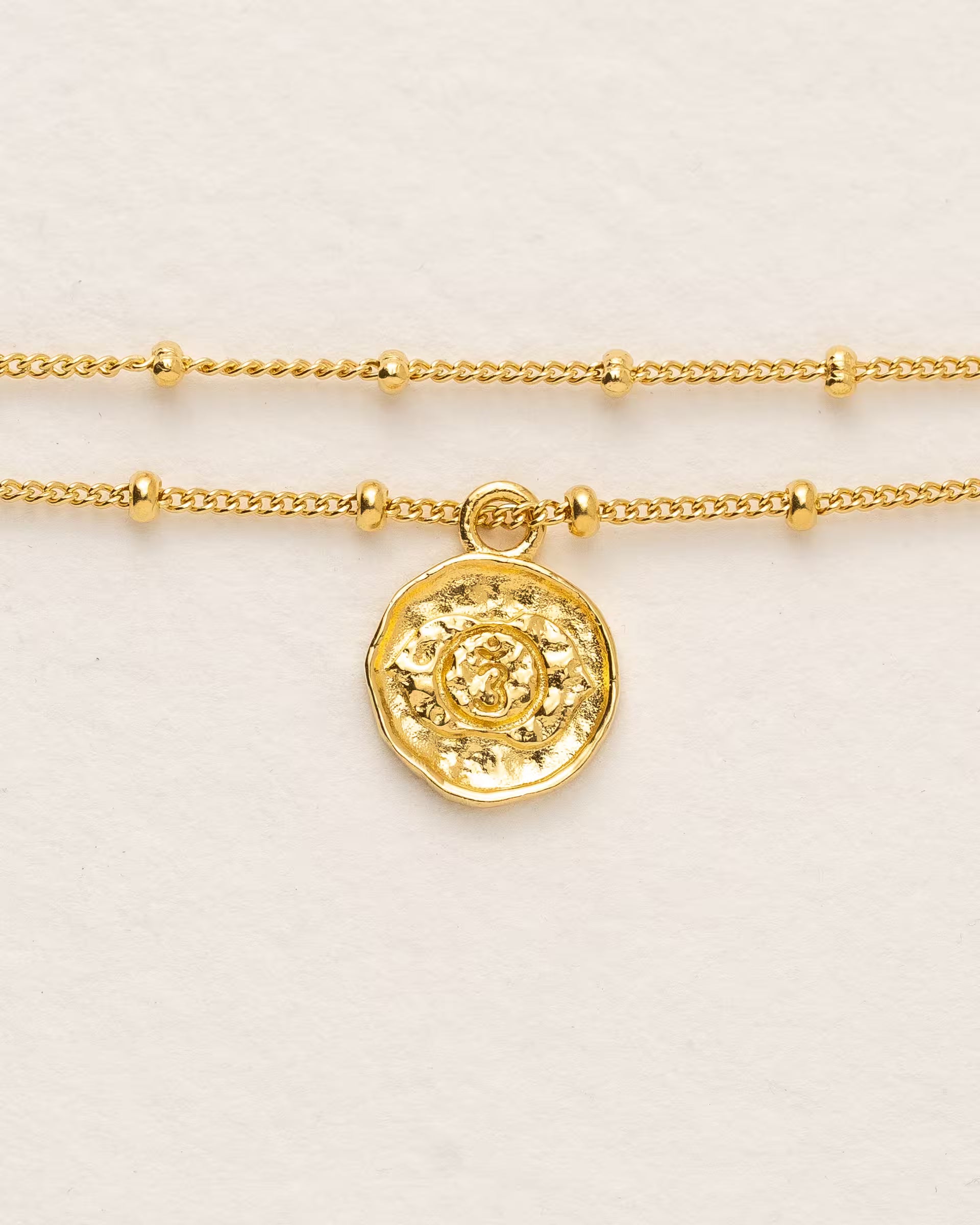 #Motiv_Ajna CHAKRA AJNA ARMBAND GOLD, goldenes Armband von ILUH, handgefertigt auf Bali – Spirituelles Produktfoto im minimalistischen Boho-Look von einem feinen, doppelten Satellitenarmband mit einem runden Münzanhänger, der das originale Sanskrit-Zeichen für das Ajna-Stirnchakra, eingebettet in kunstvolle Lotusblütenblätter zur Stärkung des Energiezentrums, zeigt.
