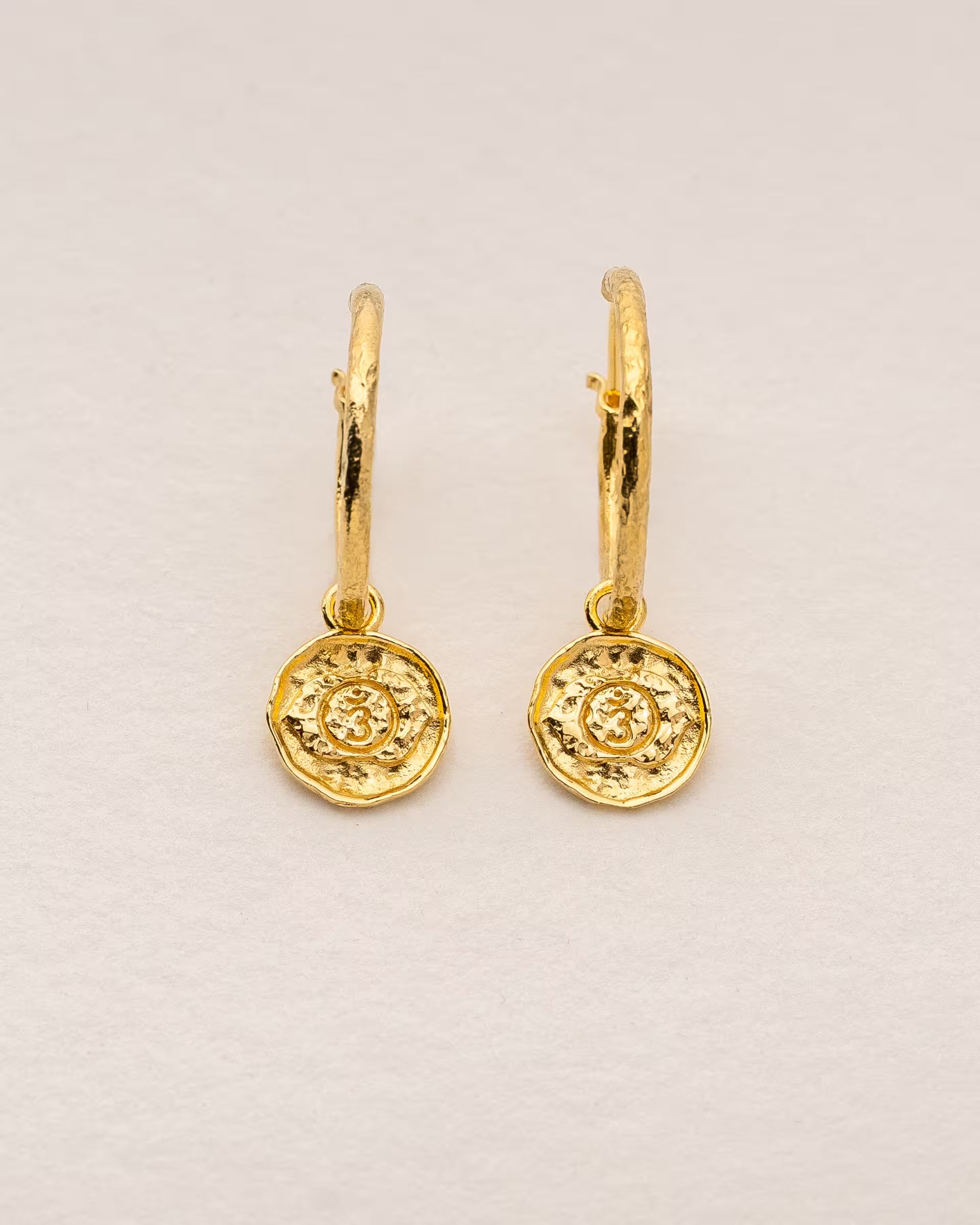 #Motiv_Ajna CHAKRA AJNA OHRRINGE GOLD, kleine goldene Creolen-Ohrringe von ILUH, handgefertigt auf Bali – Elegantes Produktfoto im spirituellen Boho-Look von einem Paar filigraner Creolen aus 18k vergoldetem 925er Silber mit je einem kleinen, runden Anhänger, der das originale Sanskrit-Zeichen für das Ajna-Stirnchakra, eingebettet in kunstvolle Lotusblütenblätter zur Stärkung des Energiezentrums, zeigt.
