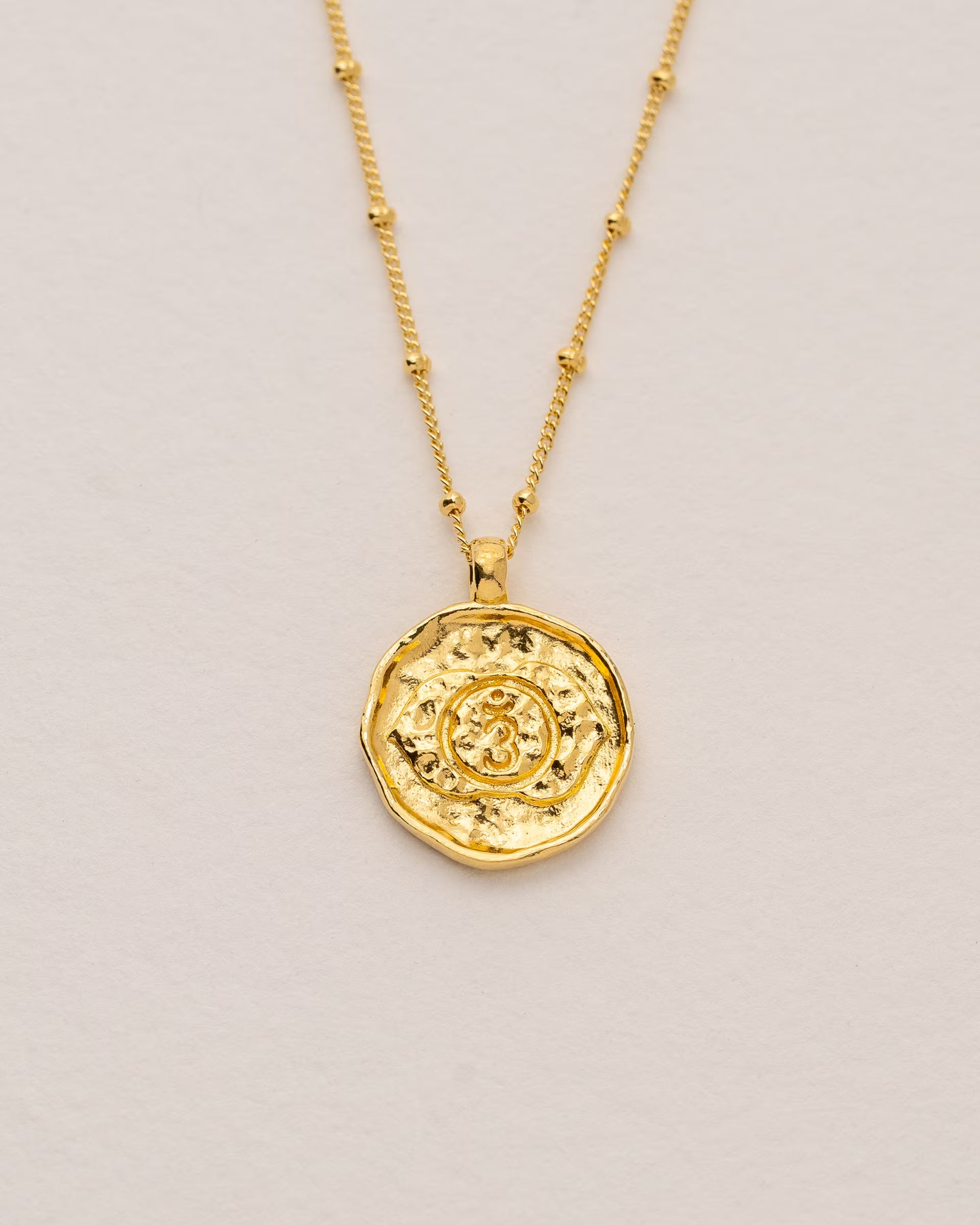 #Motiv_Ajna CHAKRA AJNA NECKLACE GOLD, goldene Halskette von ILUH, handgefertigt auf Bali – Spirituelles Produktfoto im minimalistischen Boho-Look von einer feinen Satellitenkette aus 18k vergoldetem 925er Silber mit einem runden Münzanhänger, der das detailliert geprägte Ajna-Stirnchakra-Symbol für Intuition zeigt, flach präsentiert auf einem hellen, neutralen Untergrund.