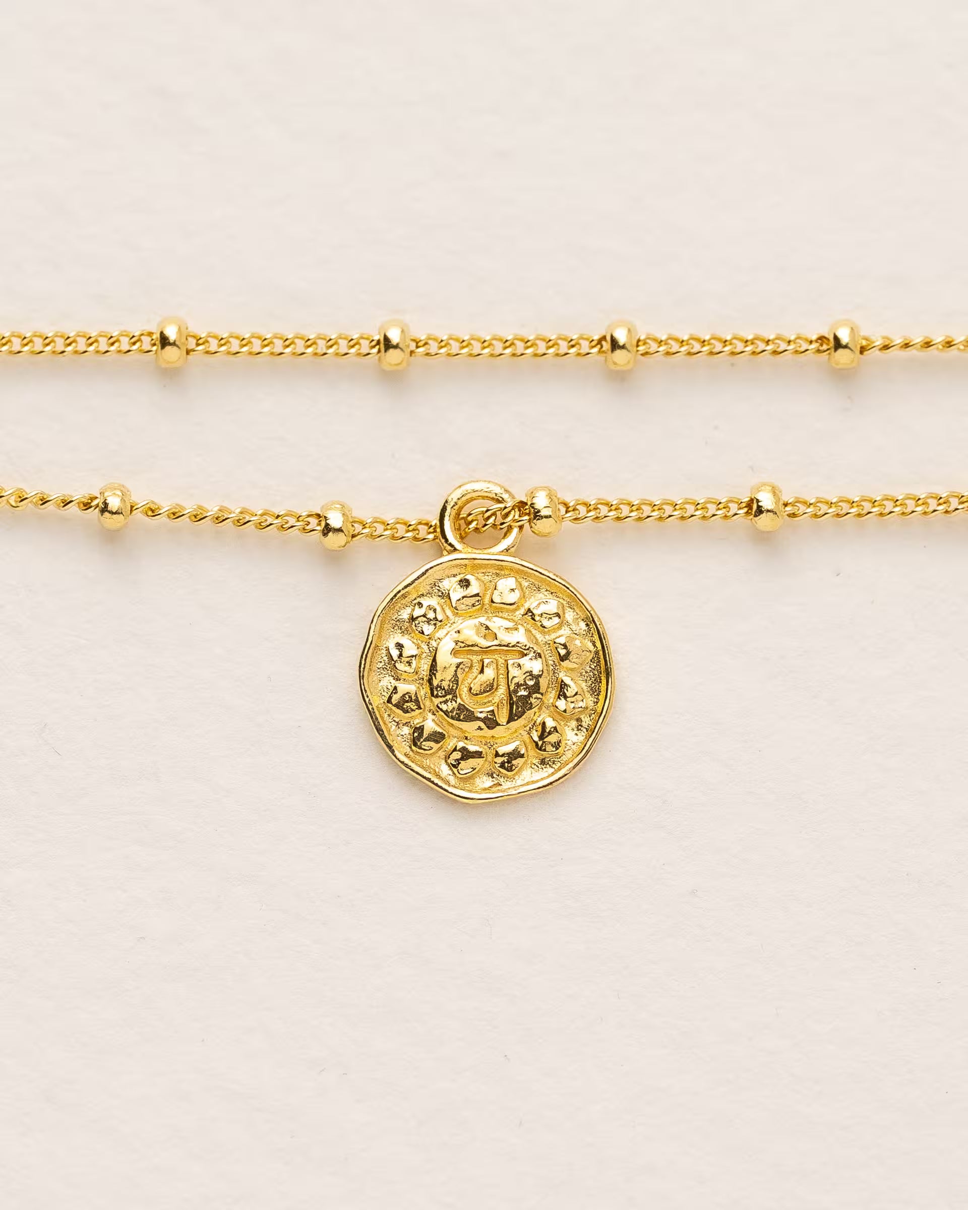 #Motiv_Anahata CHAKRA ANAHATA ARMBAND GOLD, goldenes Armband von ILUH, handgefertigt auf Bali – Spirituelles Produktfoto im minimalistischen Boho-Look von einem feinen, doppelten Satellitenarmband aus 18k vergoldetem 925er Silber mit einem runden Münzanhänger, der das originale Sanskrit-Zeichen für das Anahata-Herzchakra, eingebettet in kunstvolle Lotusblütenblätter zur Stärkung des Energiezentrums, zeigt, flach präsentiert auf einem hellen, neutralen Büttenpapier-Untergrund.