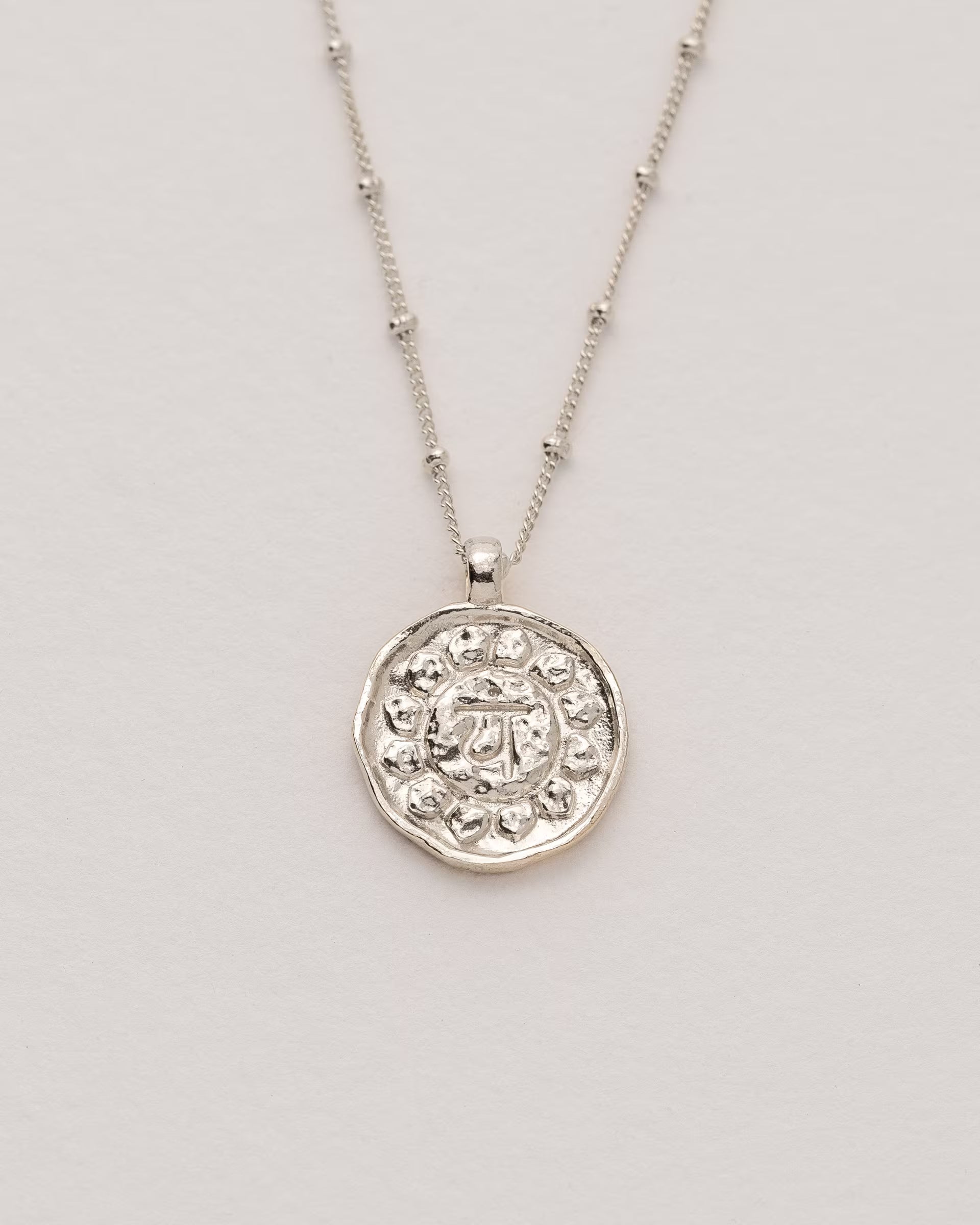 #Motiv_Anahata CHAKRA ANAHATA NECKLACE SILBER, silberne Halskette von ILUH, handgefertigt auf Bali – Spirituelles Produktfoto im minimalistischen Boho-Look von einer feinen Satellitenkette aus 925er Silber mit einem runden Münzanhänger, der das originale Sanskrit-Zeichen für das Anahata-Herzchakra eingebettet in kunstvolle Lotusblütenblätter zur Stärkung des Energiezentrums zeigt, flach präsentiert auf einem hellen, neutralen Büttenpapier-Untergrund.