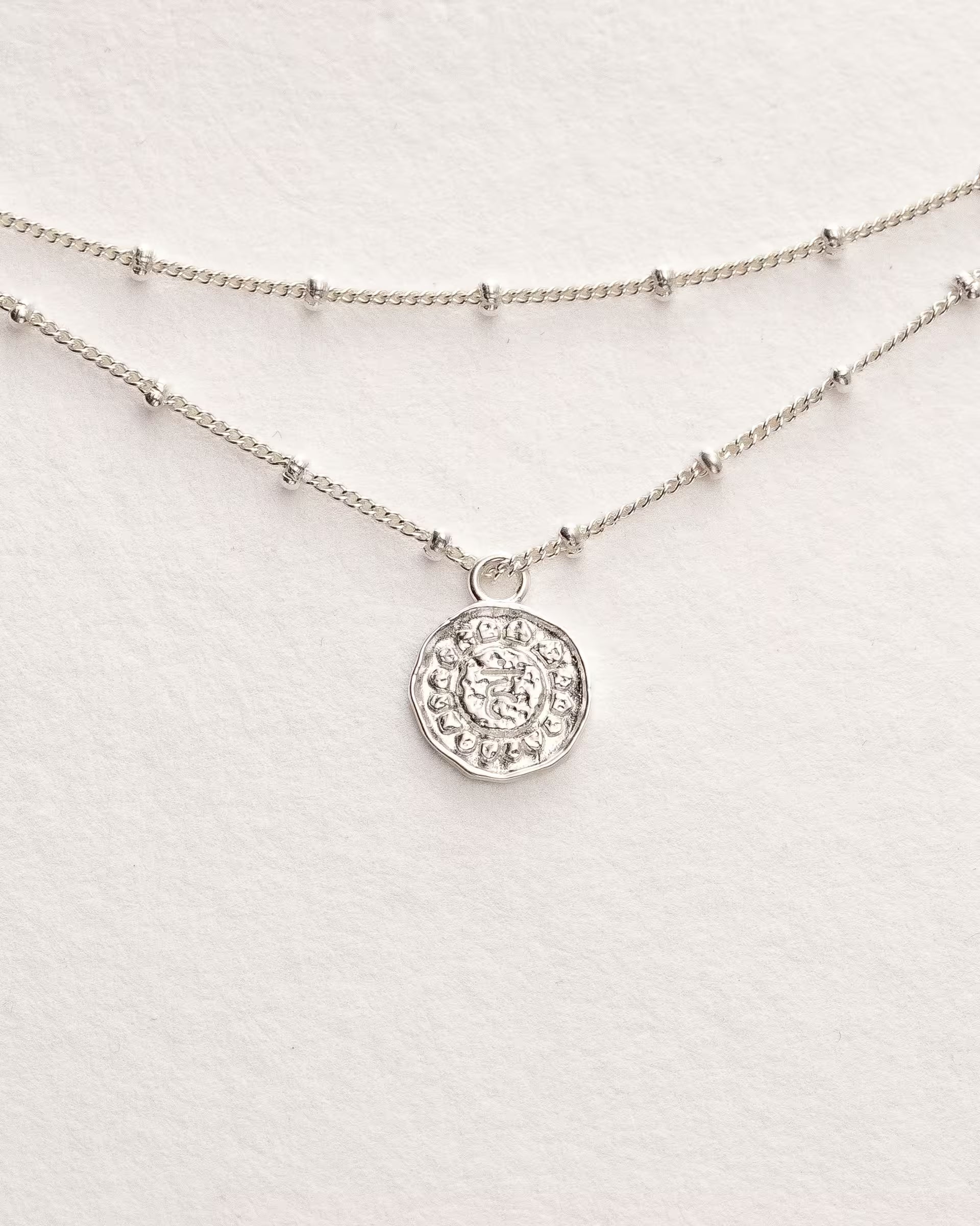 Filigranes ILUH Chakra-Armband in 925er Sterlingsilber mit geprägtem Münz-Anhänger und feiner, doppelter Kugelkette; das Design verkörpert achtsame Eleganz und moderne Spiritualität. Präsentiert auf hochwertigem Büttenpapier für einen minimalistischen, femininen Look.