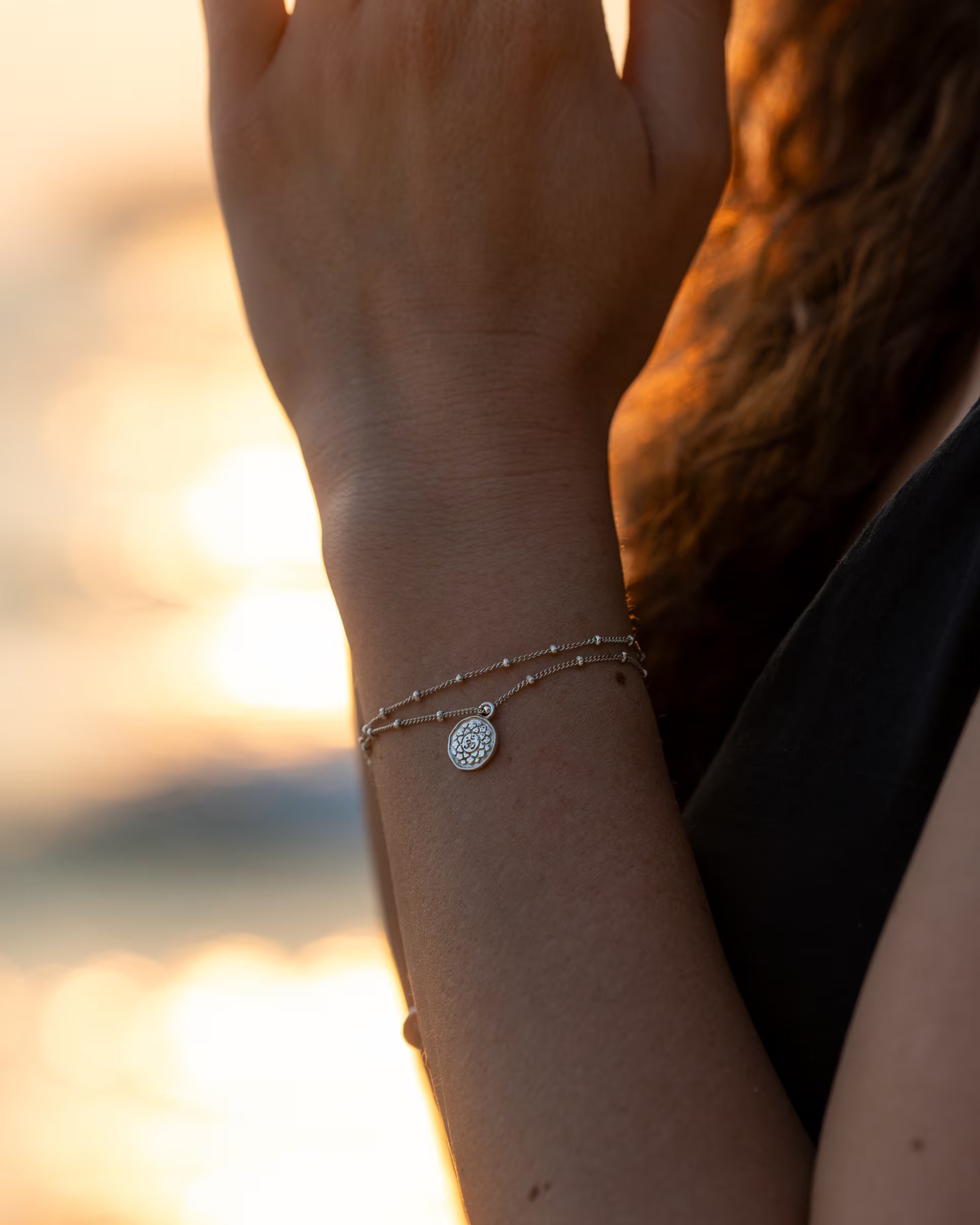 CHAKRA ARMBAND SILBER, silberner Schmuck von ILUH, handgefertigt auf Bali – Sinnliche Nahaufnahme am sommerlichen Strand von dem Handgelenk einer Frau mit zum Gebet gefalteten Händen, die das filigrane, zweireihige Chakra-Armband an einer feinen Satellitenkette mit rundem Motiv-Anhänger im warmen Gegenlicht des Sonnenuntergangs zeigt.