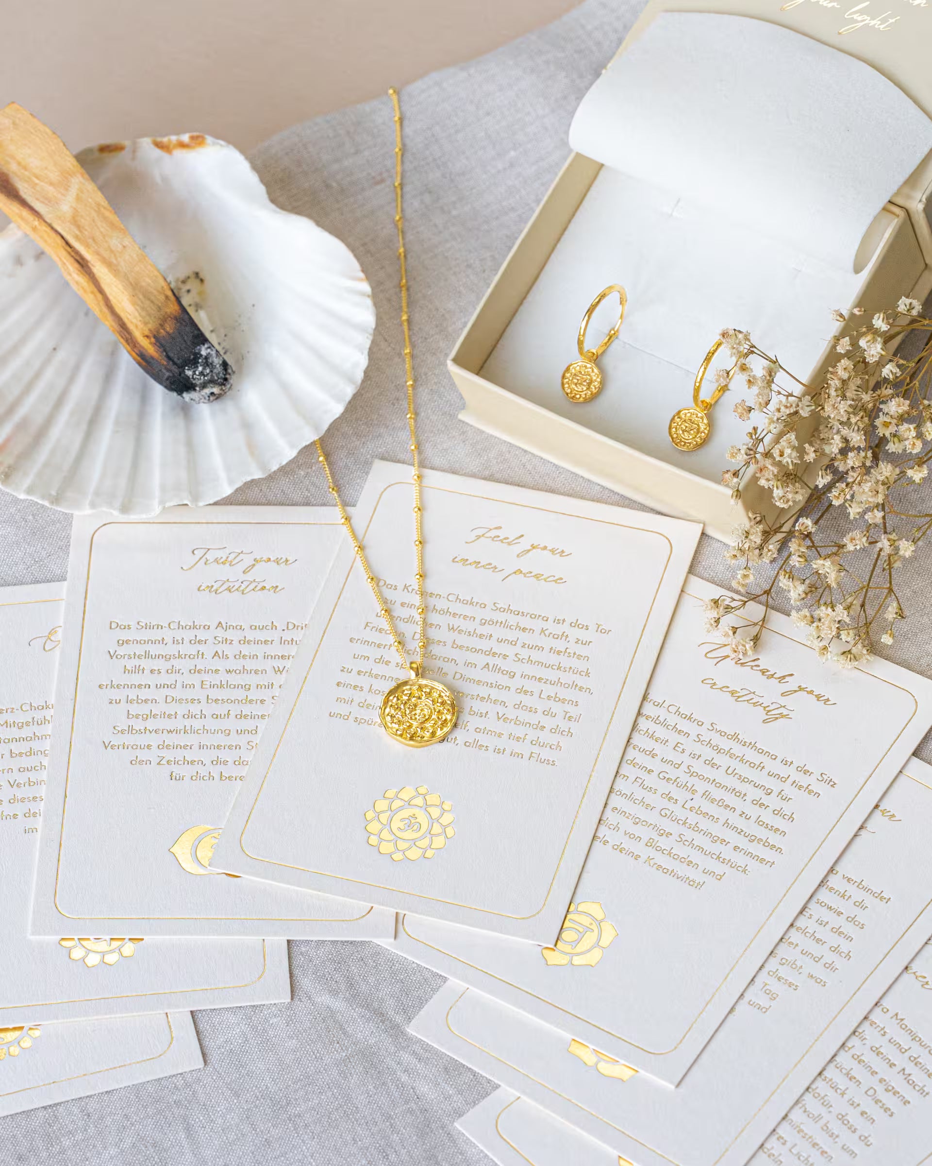 Goldene Chakra-Halskette und Ohrringe aus dem Schmuck-Label ILUH, präsentiert auf ästhetischen Intentionskarten mit Palo Santo und Schleierkraut, die die spirituelle Bedeutung der Symbole erklären; das Bild vermittelt den ganzheitlichen Ansatz der auf Bali handgefertigten Kollektion.
