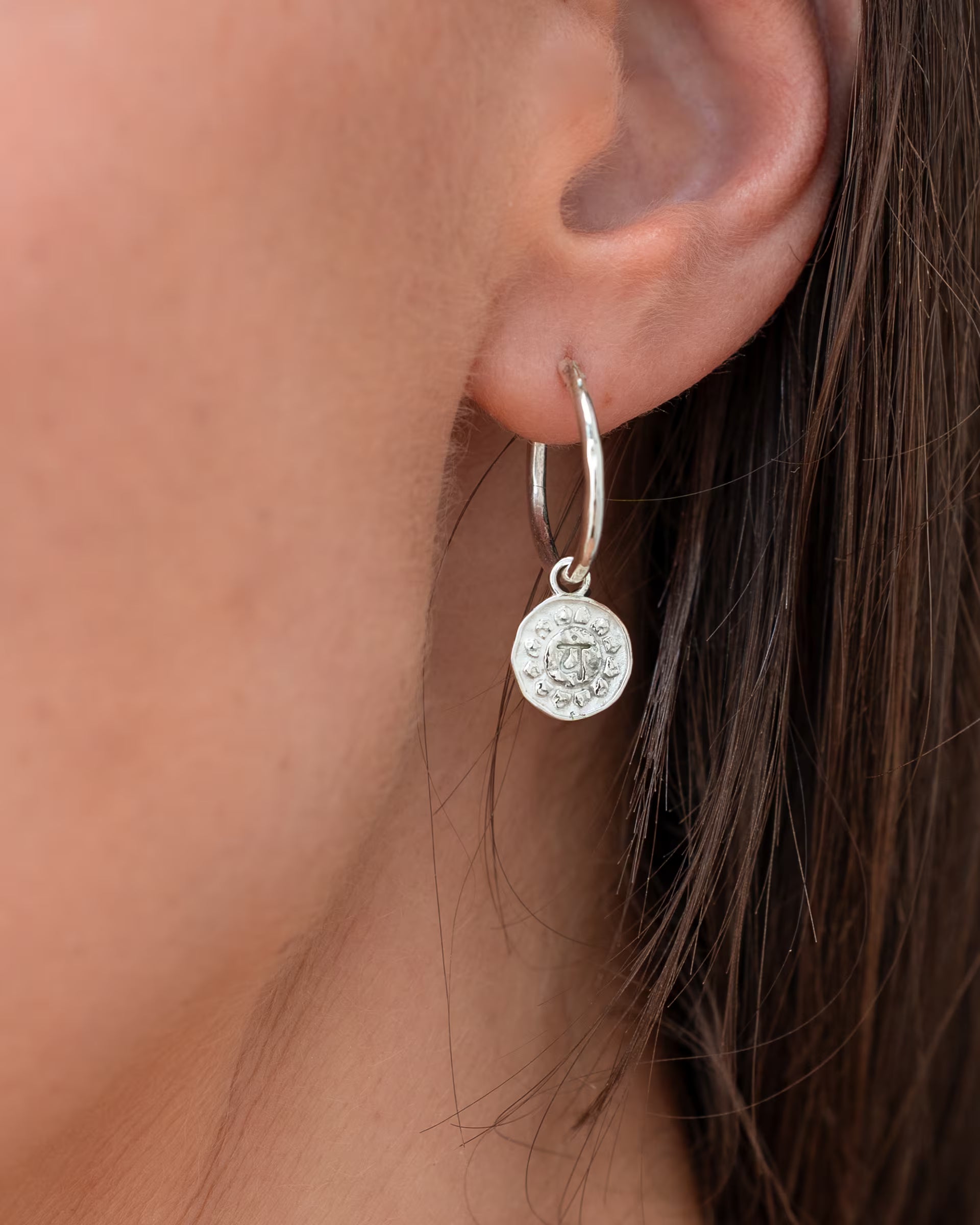 CHAKRA OHRRINGE SILBER, silberner Schmuck von ILUH, handgefertigt auf Bali – Detailreiche Nahaufnahme im Profil von dem Ohr einer Frau mit braunen Haaren, die eine filigrane, mittelgroße Creole aus 925er Silber mit einem runden, fein geprägten Chakra-Münzanhänger präsentiert.