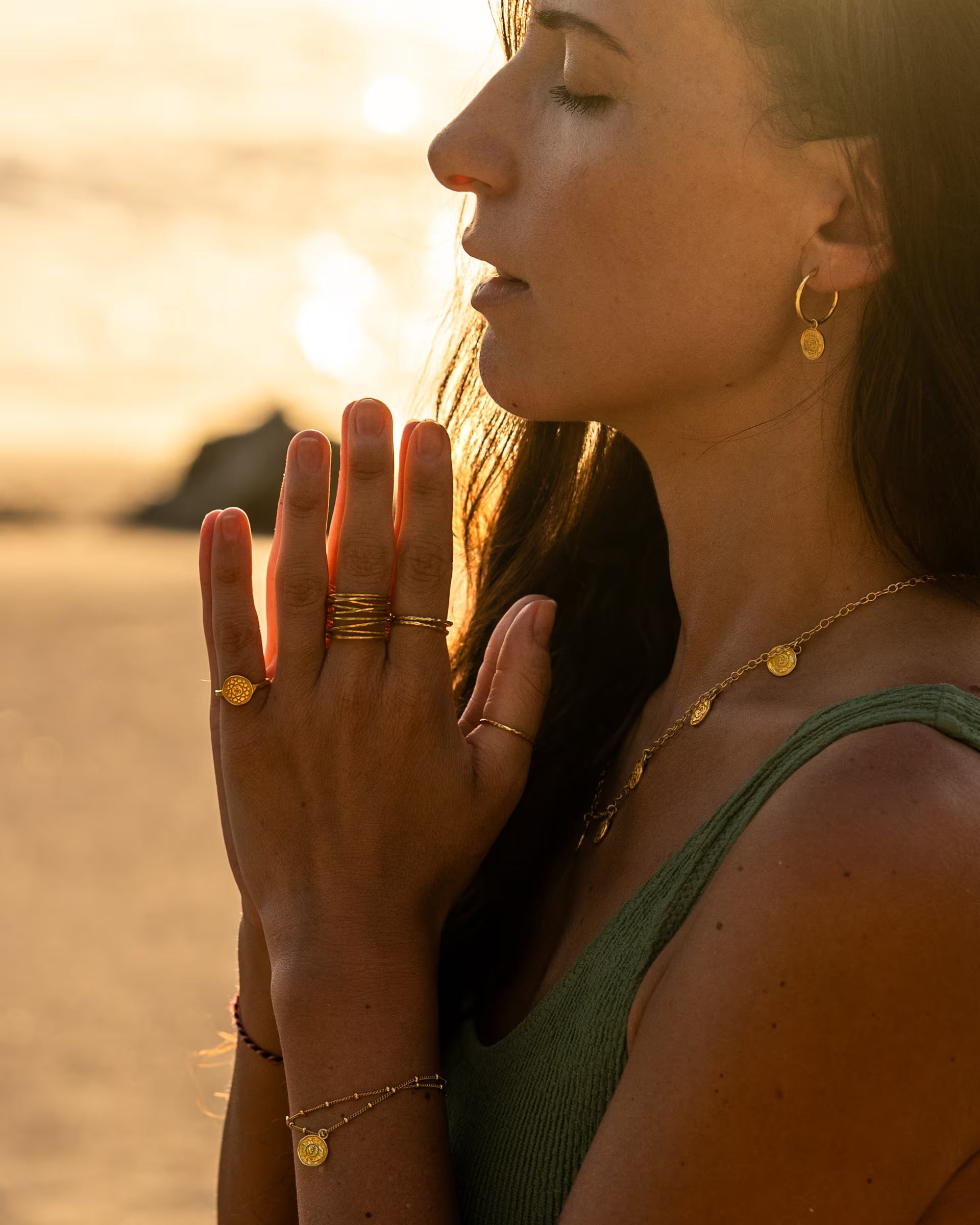 CHAKRA SCHMUCKSET, goldene Halskette, Ohrringe und Ringe von der Schmuckmarke ILUH, handgefertigt auf Bali – Spirituelle Nahaufnahme im Profil von einer jungen Frau mit geschlossenen Augen im salbeigrünen Oberteil, die am Strand die Hände zum Gebet faltet und dabei die feine PRANA Plättchenkette, hängende Chakra-Creolen, das zarte Armband sowie mehrere stacking Ringe aus 18k vergoldetem 925er Silber im warmen Gegenlicht zeigt.