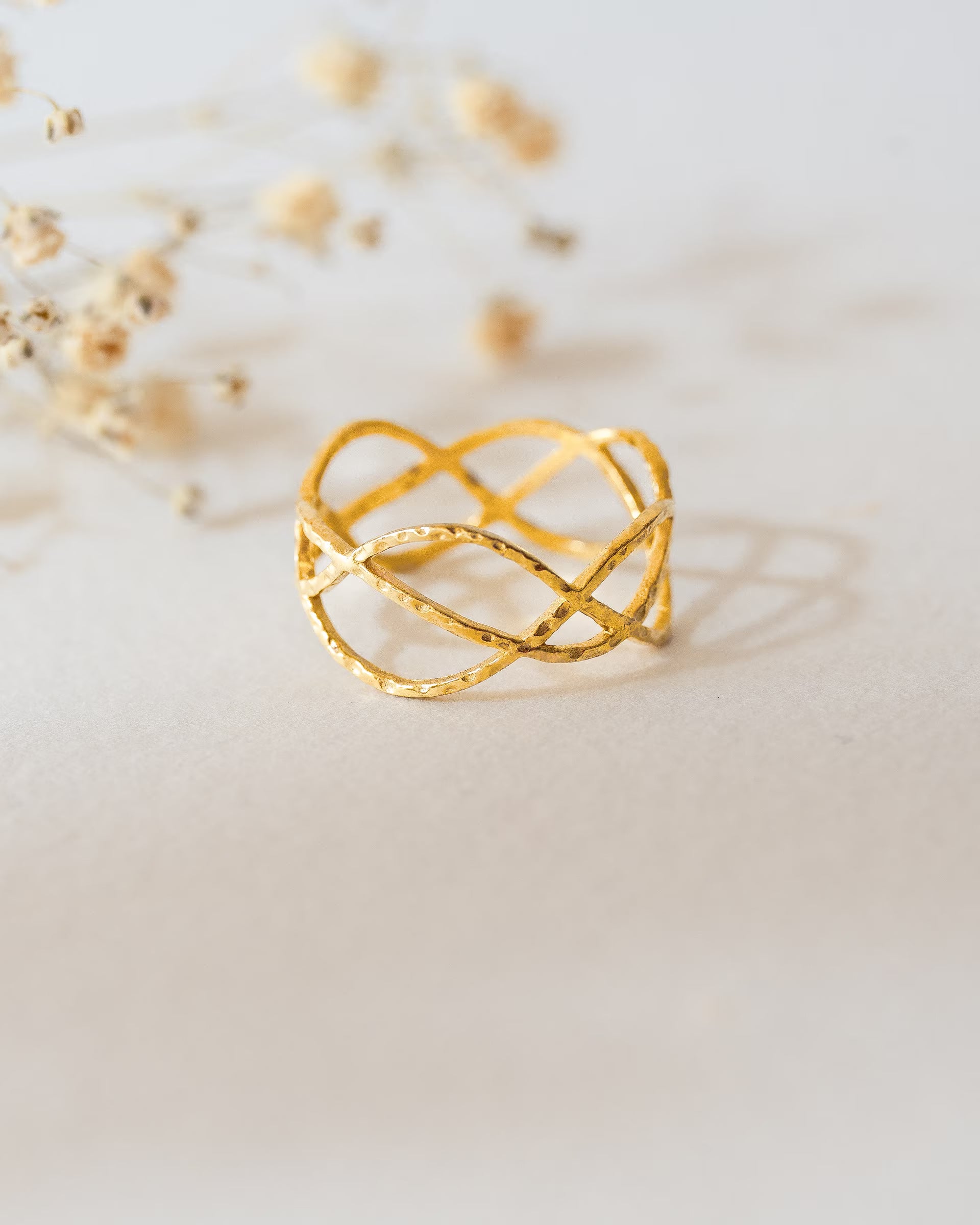 CHAKRA LOTUS RING GOLD, breiter goldener Ring von ILUH, handgefertigt auf Bali – Spirituelles Produktfoto im zarten Boho-Look von einem breiten Ring aus 18k vergoldetem 925er Silber mit einem filigranen, organisch geschwungenen und gehämmerten Wellen-Muster, das an eine abstrakte Lotusblüte erinnert, präsentiert auf einem hellen Untergrund vor dezenten Trockenblumen.