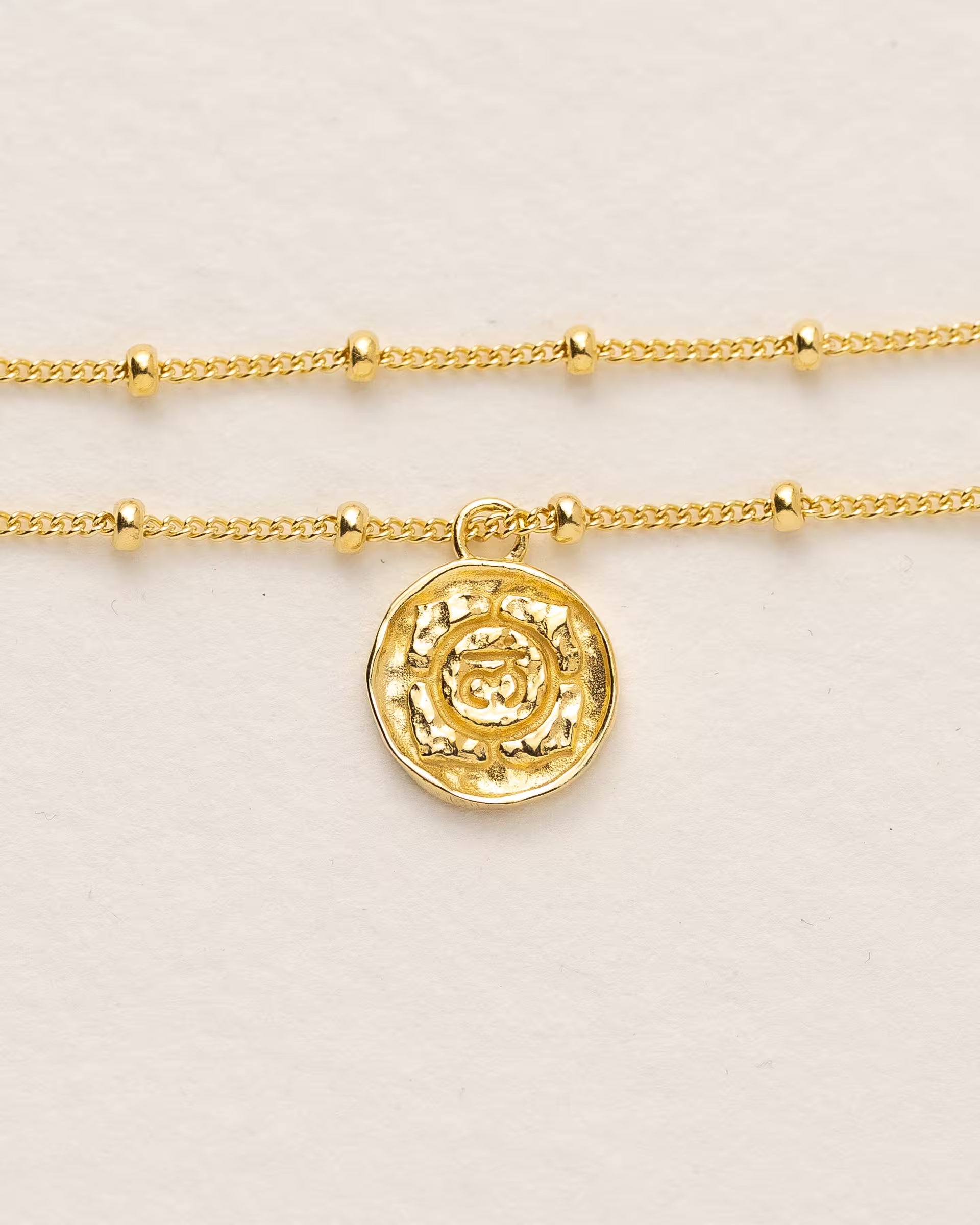 #Motiv_Muladhara CHAKRA MULADHARA ARMBAND GOLD, goldenes Armband von ILUH, handgefertigt auf Bali – Spirituelles Produktfoto im minimalistischen Boho-Look von einem feinen, doppelten Satellitenarmband mit einem runden Münzanhänger, der das originale Sanskrit-Zeichen für das Muladhara-Wurzelchakra, eingebettet in kunstvolle Lotusblütenblätter zur Stärkung des Energiezentrums, zeigt.
