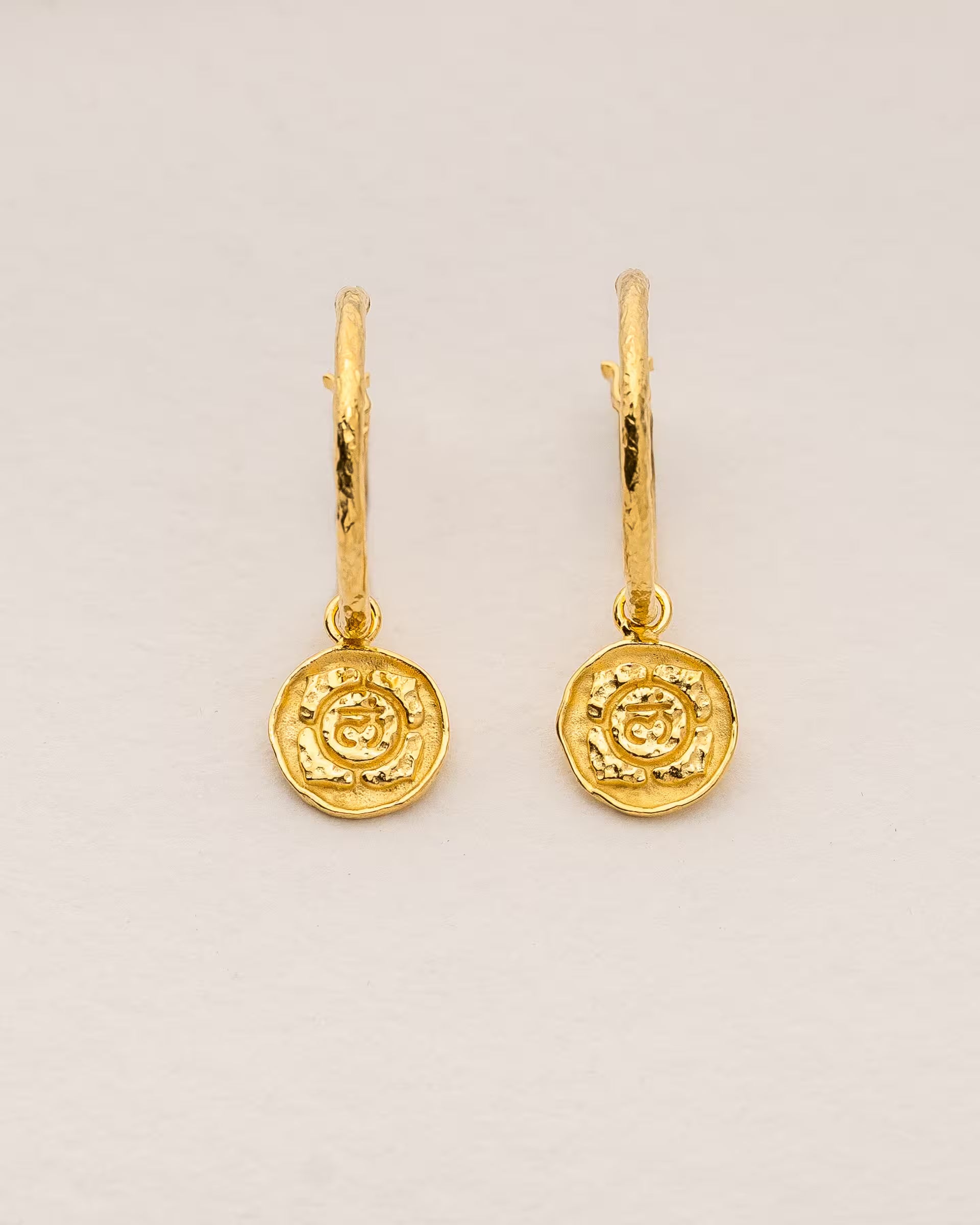 #Motiv_Muladhara CHAKRA MULADHARA OHRRINGE GOLD Produktbild – Diese handgefertigten Ohrringe von ILUH aus Bali im minimalistischen Boho-Stil bestehen aus strukturierten Creolen mit einem runden Anhänger, der das originale Sanskrit-Zeichen für das Muladhara-Wurzelchakra zeigt; eingebettet in kunstvolle Lotusblütenblätter dienen sie als kraftvolles Symbol zur Stärkung deines Energiezentrums.