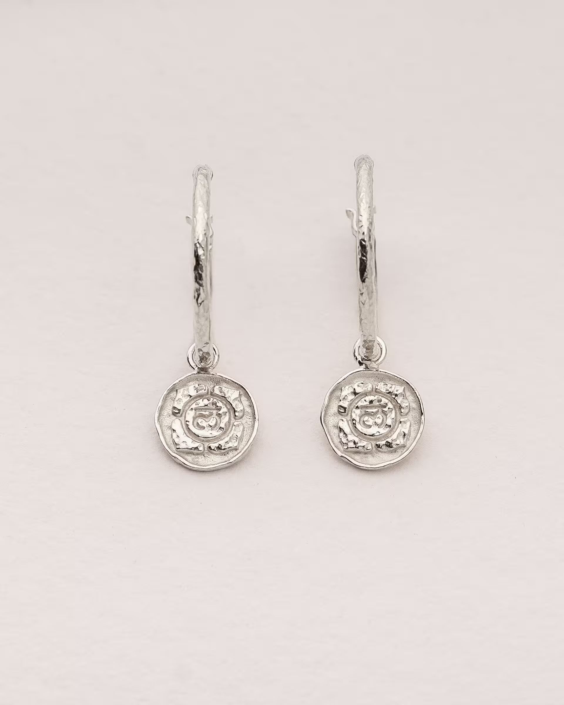 #Motiv_Muladhara CHAKRA MULADHARA OHRRINGE SILBER Produktbild – Diese handgefertigten Ohrringe von ILUH aus Bali im minimalistischen Boho-Stil bestehen aus strukturierten Creolen mit einem runden Anhänger, der das originale Sanskrit-Zeichen für das Muladhara-Wurzelchakra zeigt; eingebettet in kunstvolle Lotusblütenblätter dienen sie als kraftvolles Symbol zur Stärkung deines Energiezentrums.