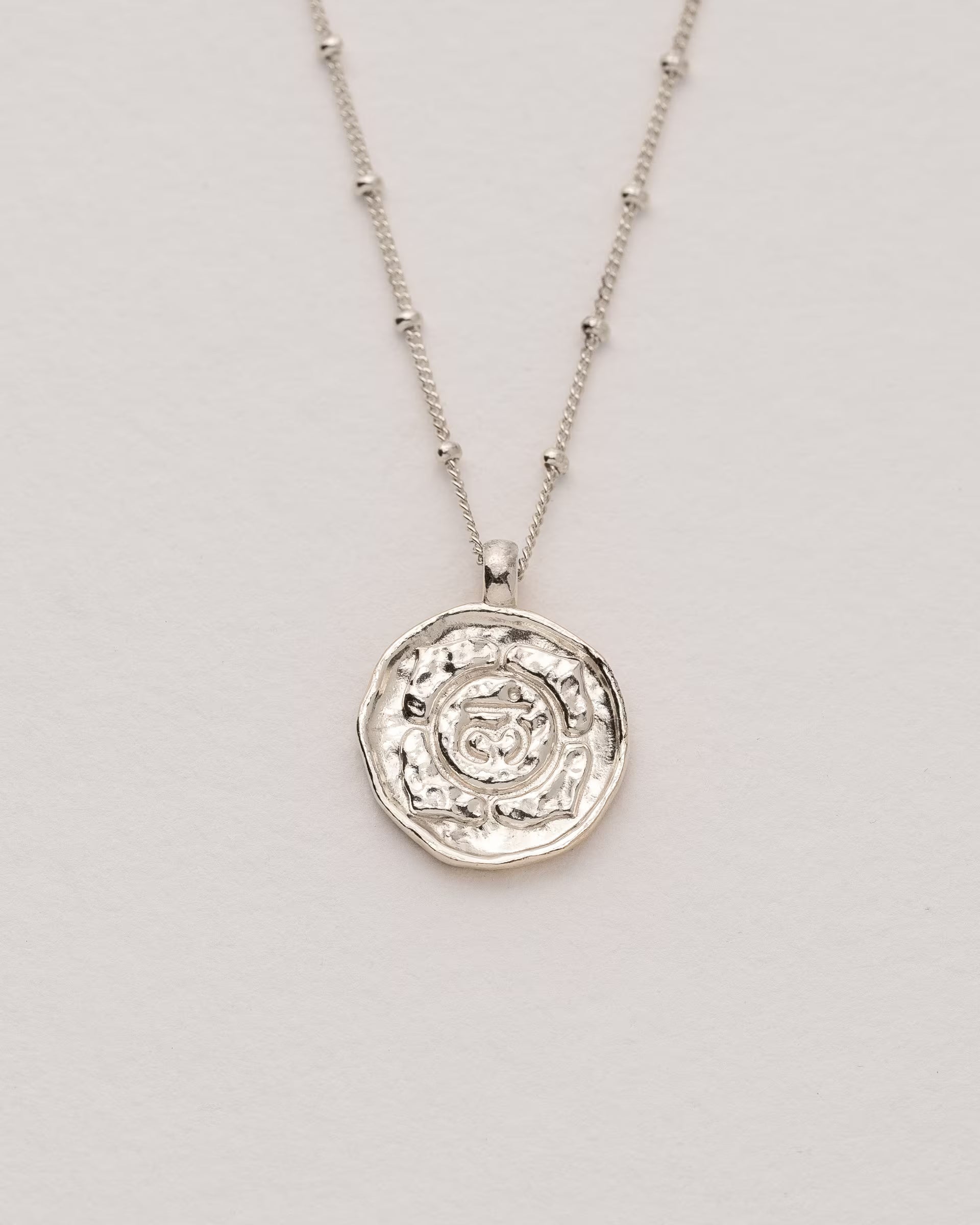 #Motiv_Muladhara CHAKRA MULADHARA NECKLACE SILBER, silberne Halskette von ILUH, handgefertigt auf Bali – Spirituelles Produktfoto im minimalistischen Boho-Look von einer feinen Satellitenkette aus 925er Silber mit einem runden Münzanhänger, der das originale Sanskrit-Zeichen für das Muladhara-Wurzelchakra, eingebettet in kunstvolle Lotusblütenblätter zur Stärkung des Energiezentrums, zeigt.