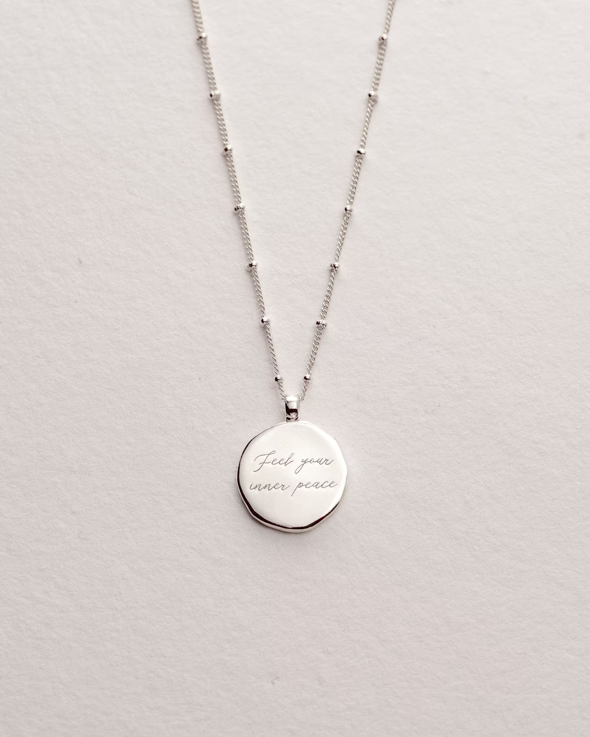 Filigrane ILUH Saturn Chain aus 925er Sterlingsilber mit feinen Kugel-Elementen und einem runden, handgefertigten Anhänger; die Gravur ‚Feel your inner peace‘ symbolisiert das Sahasrara-Chakra (Kronenchakra) und steht für universelle Verbundenheit und spirituellen Frieden. Präsentiert auf strukturiertem Büttenpapier für einen modernen, minimalistischen und femininen Look.
