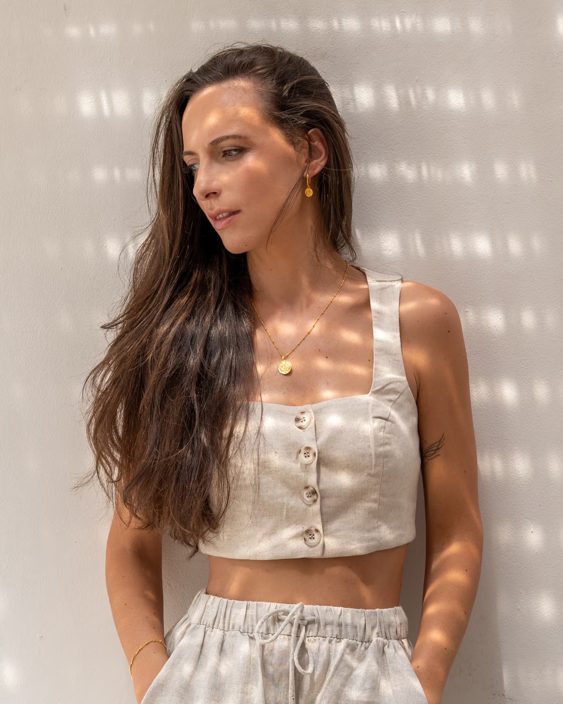 Elegante Nahaufnahme im sommerlichen Look von einer wegschauenden Frau mit langen, braunen Haaren in einem hellen Leinen-Zweiteiler vor einer weißen Wand mit sanftem Licht- und Schattenspiel, die eine Kombination aus auf Bali handgefertigtem goldenem Schmuck aus der Chakra-Kollektion – bestehend aus der CHAKRA KETTE mit rundem Münzanhänger und den passenden CHAKRA OHRRINGEN – aus 18k vergoldetem 925er Silber von der Schmuckmarke ILUH trägt.