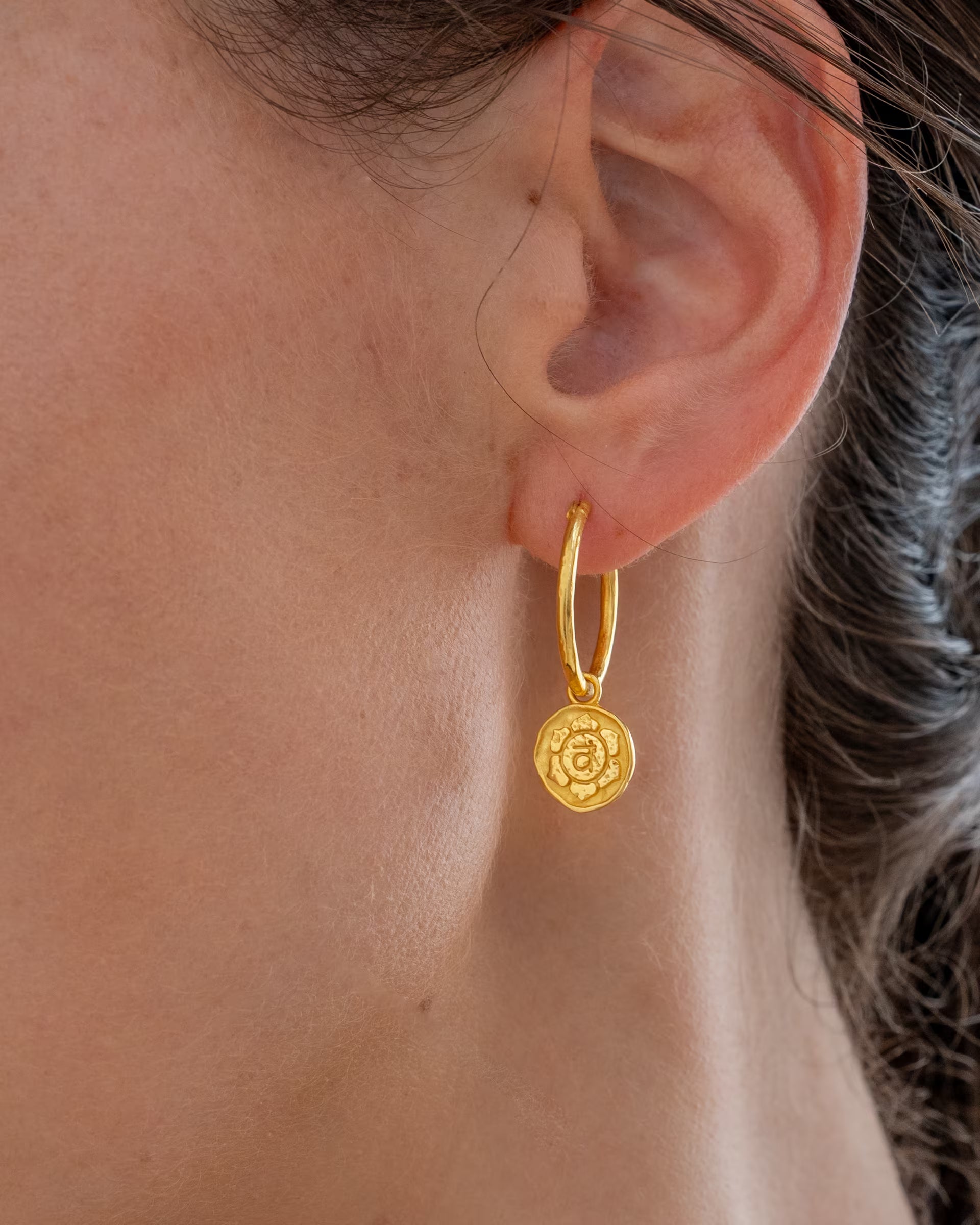 CHAKRA OHRRINGE GOLD, goldener Schmuck von ILUH, handgefertigt auf Bali – Detailreiche Nahaufnahme im Profil von dem Ohr einer Frau, die eine elegante, gehämmerte, mittelgroße Creole aus 18k vergoldetem 925er Silber mit einem runden, fein geprägten Chakra-Münzanhänger präsentiert.
