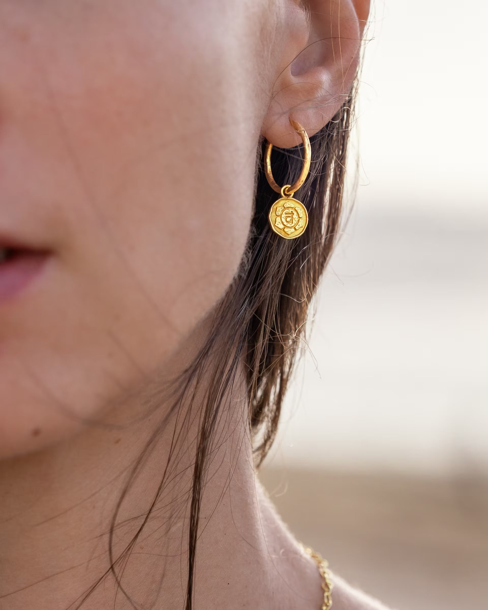 CHAKRA OHRRINGE GOLD, goldener Schmuck von ILUH, handgefertigt auf Bali – Detailreiche Nahaufnahme im Profil von dem Gesicht einer Frau, die eine filigrane gehämmerte Creole aus 18k vergoldetem 925er Silber mit einem runden, detailliert geprägten Chakra-Anhänger am Meer präsentiert.