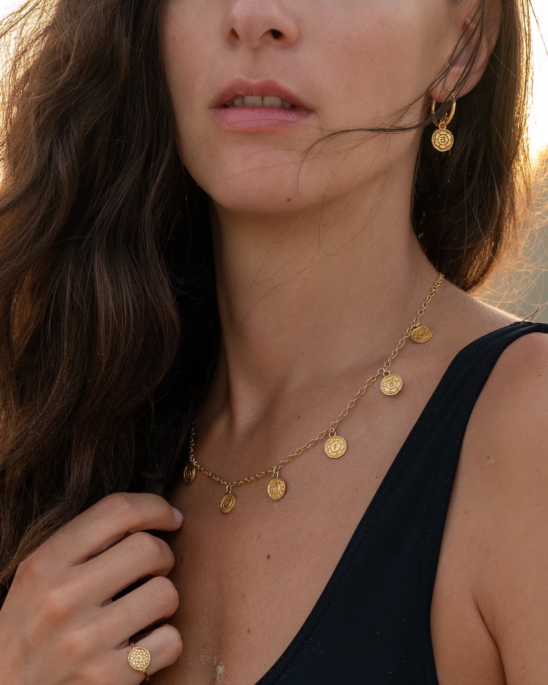 CHAKRA SCHMUCKSET, goldene Halskette und Creolen von ILUH, handgefertigt auf Bali – Detailreiche Nahaufnahme im spirituellen Beach-Look von einer Frau im schwarzen Badeanzug, die im warmen Licht des Sonnenuntergangs die PRANA KETTE mit sieben runden Chakra-Münzanhängern, die passenden CHAKRA OHRRINGE als Creolen sowie einen geprägten Chakra Motiv-Ring aus 18k vergoldetem 925er Silber zeigt.