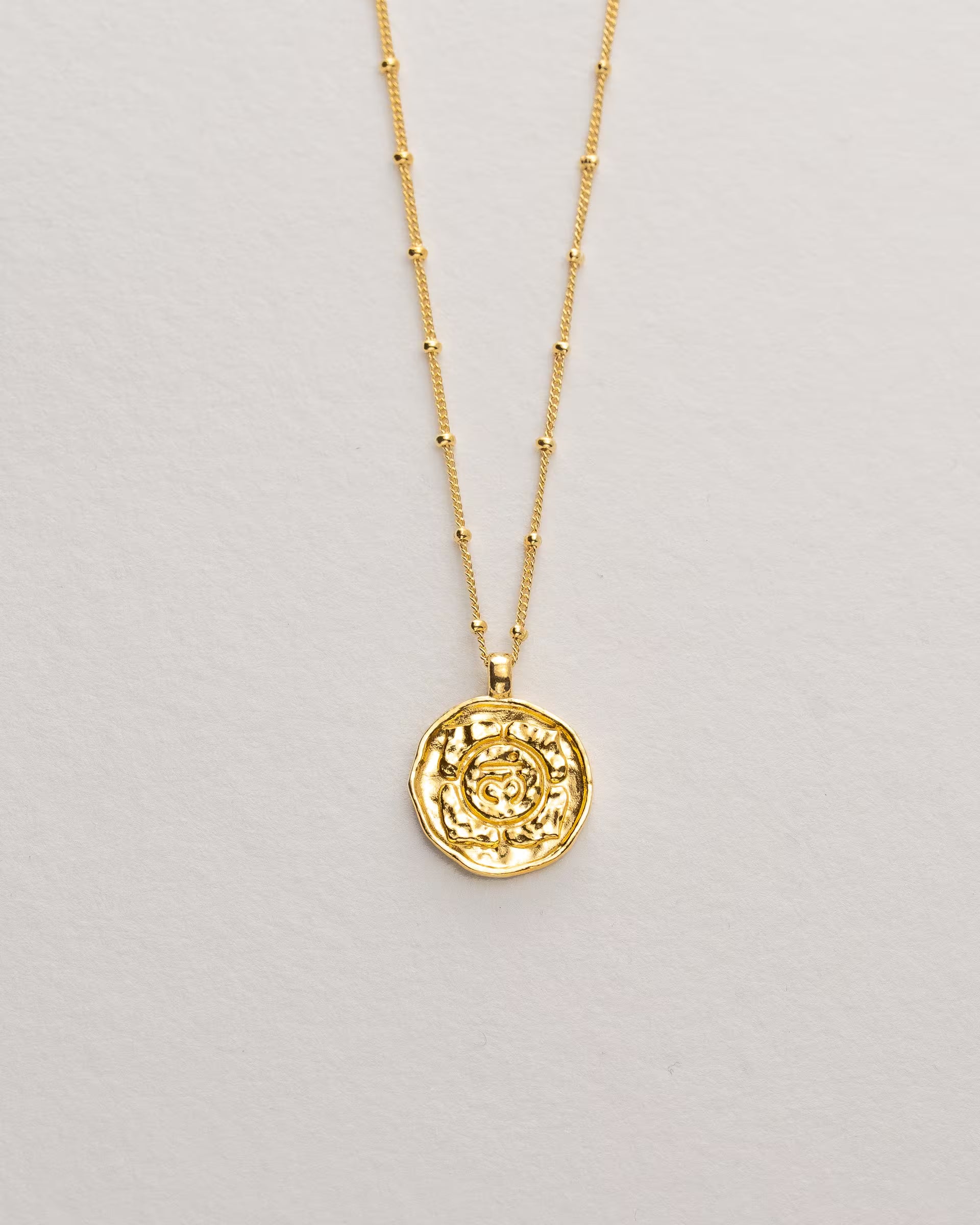 Filigrane ILUH Halskette aus 18k vergoldetem, recyceltem Silber mit einer Saturn Kette und einem kreisrunden Anhänger, der ein geprägtes Kronen-Chakra-Symbol zeigt; Nahaufnahme auf strukturiertem Hintergrund, die das handgefertigte Design und die sanfte Ästhetik für spirituellen, modernen Schmuck hervorhebt.