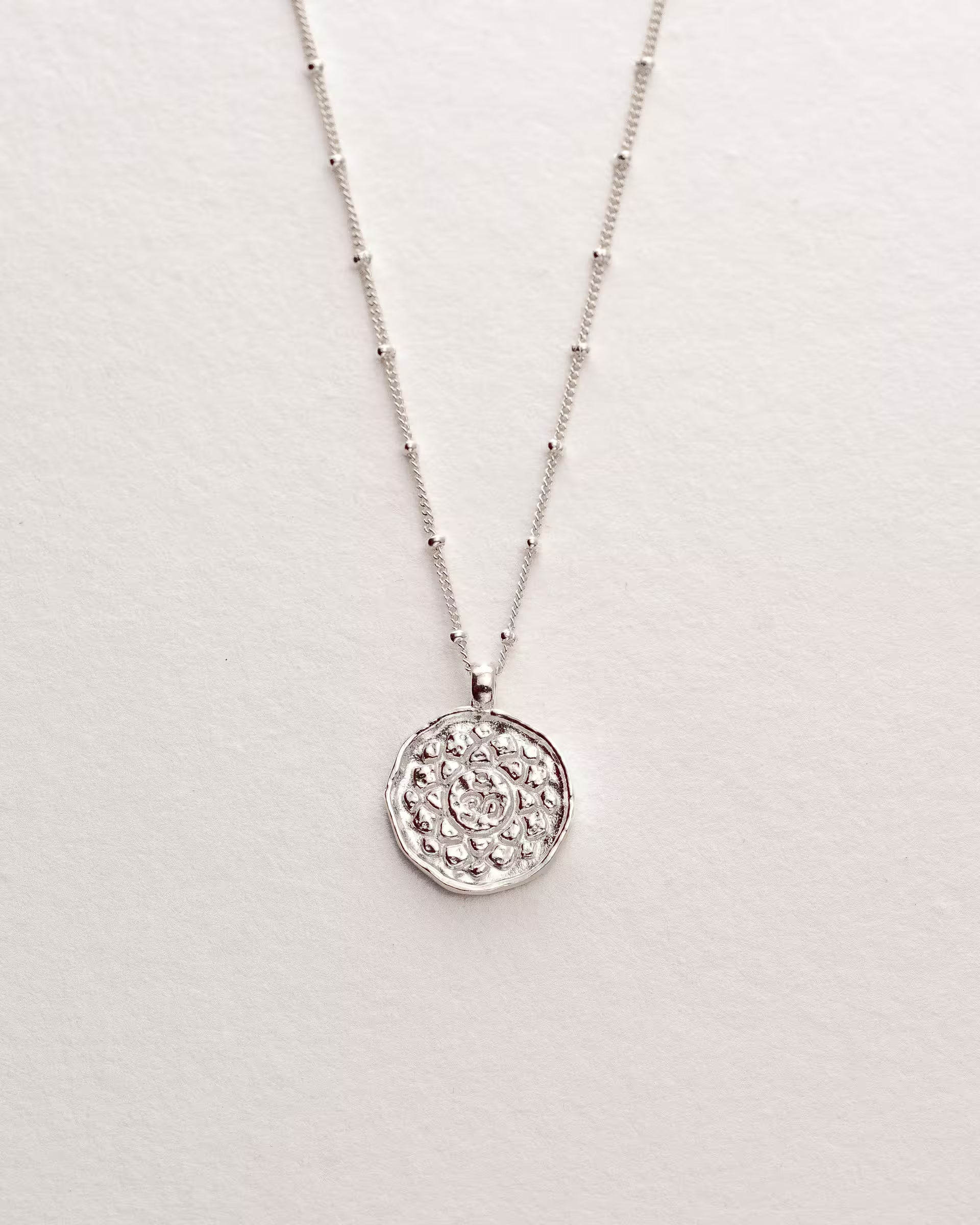 Filigrane ILUH Halskette aus 925er Sterlingsilber mit einer feinen Kugelketten-Struktur und einem runden, geprägten Anhänger; Nahaufnahme auf strukturiertem Büttenpapier, die das moderne, handgefertigte Design als spirituelles Schmuckstück für Klarheit und Intuition hervorhebt.