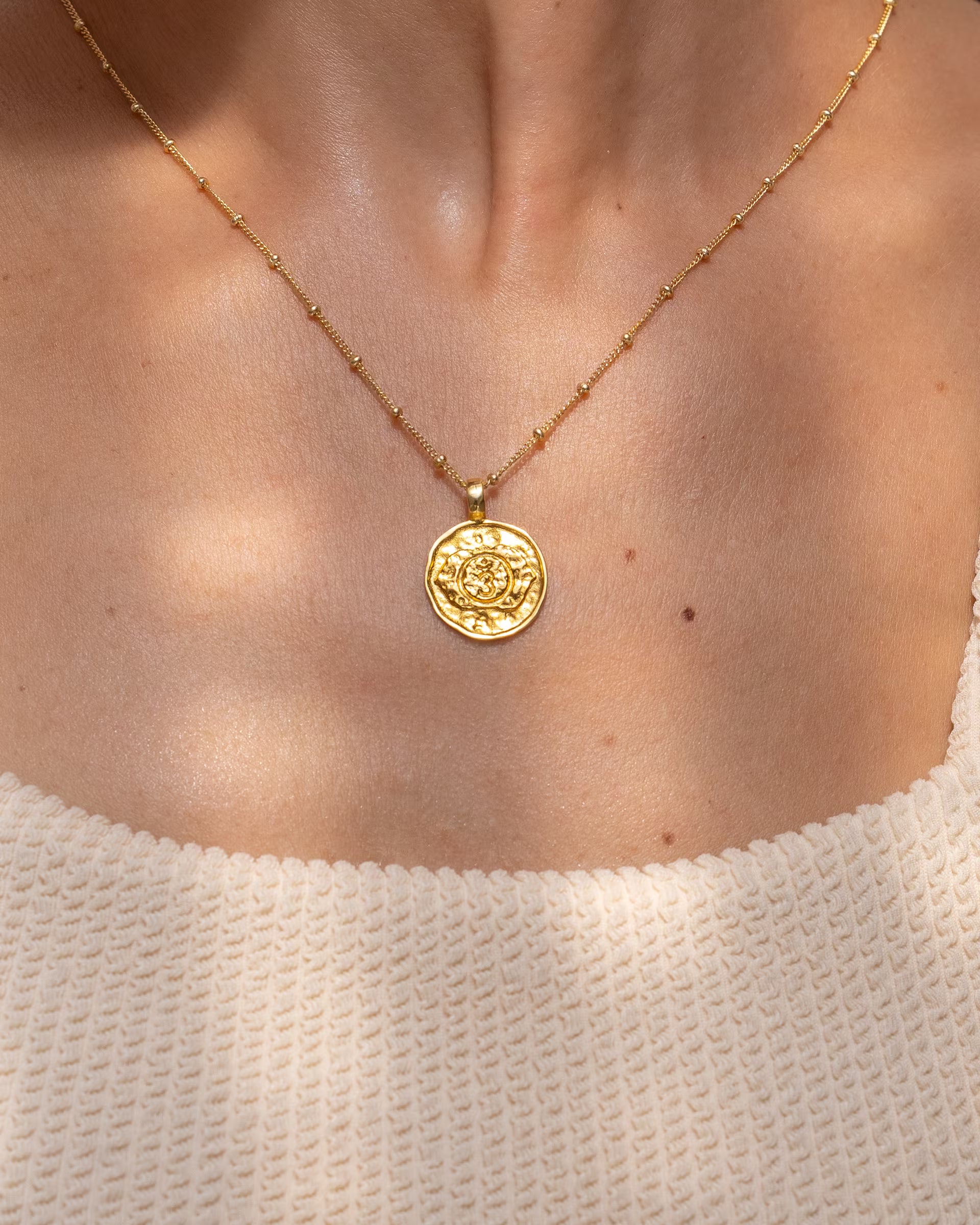 Nahaufnahme im minimalistisch-eleganten Look von dem Dekolletee einer Frau im hellen Rippstrick-Oberteil, die die auf Bali handgefertigte goldene CHAKRA KETTE mit einem runden, detailreich geprägten Chakra-Münzanhänger an einer feinen Kugelkette (Satellitenkette) aus 18k vergoldetem 925er Silber von der Schmuckmarke ILUH trägt.