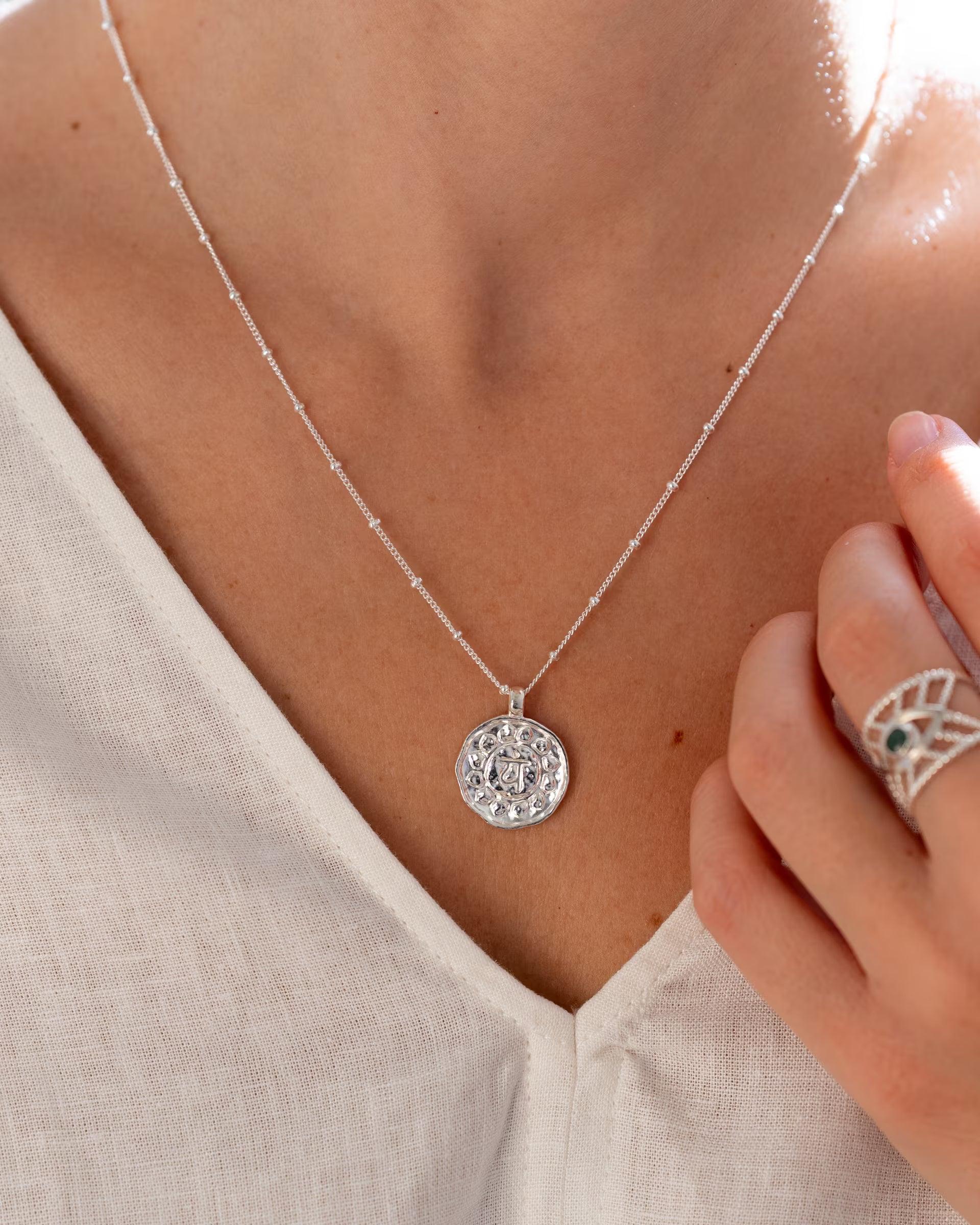 Nahaufnahme im minimalistisch-eleganten Look von dem Dekolletee einer Frau im hellen Leinen-Oberteil, die die auf Bali handgefertigte silberne CHAKRA KETTE mit einem runden, detailreich geprägten Chakra-Münzanhänger an einer feinen Satellitenkette sowie den breiten INTUITION RING mit grünem Smaragd Edelstein aus 925er Silber von der Schmuckmarke ILUH trägt.