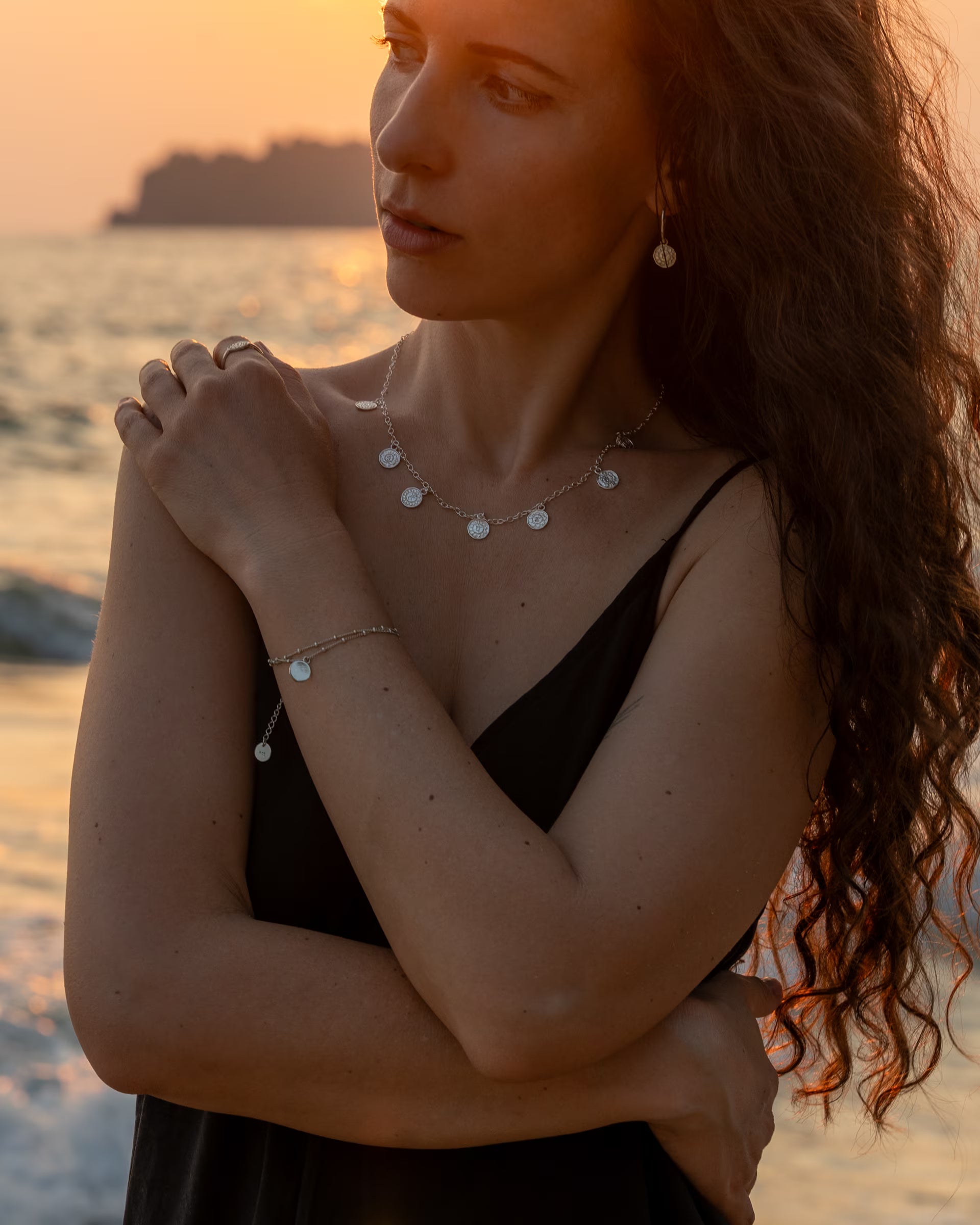 Handgefertigte PRANA Kette mit allen 7 Chakren-Motiven aus 925er Silber von der Schmuckmarke ILUH, kombiniert mit Chakra-Armband, Chakra-Ring und Chakra Creolen-Ohrringen als silbernes Schmuck-Stacking, getragen von einer Frau mit langen Haaren im eleganten Strand-Look am Meer im warmen Abendlicht.