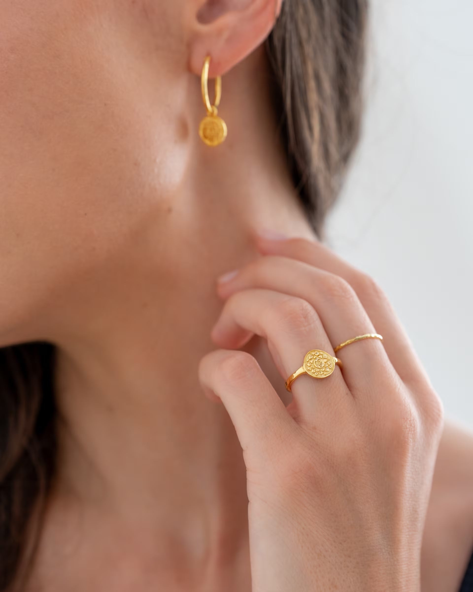CHAKRA RING UND OHRRINGE, goldener Schmuck von ILUH, handgefertigt auf Bali – Elegante Nahaufnahme von einer Frau, die eine Hand zart an ihren Hals legt und dabei den runden, fein geprägten CHAKRA RING am Zeigefinger sowie die passenden hängenden CHAKRA OHRRINGE als kleine Creolen aus 18k vergoldetem 925er Silber zeigt.
